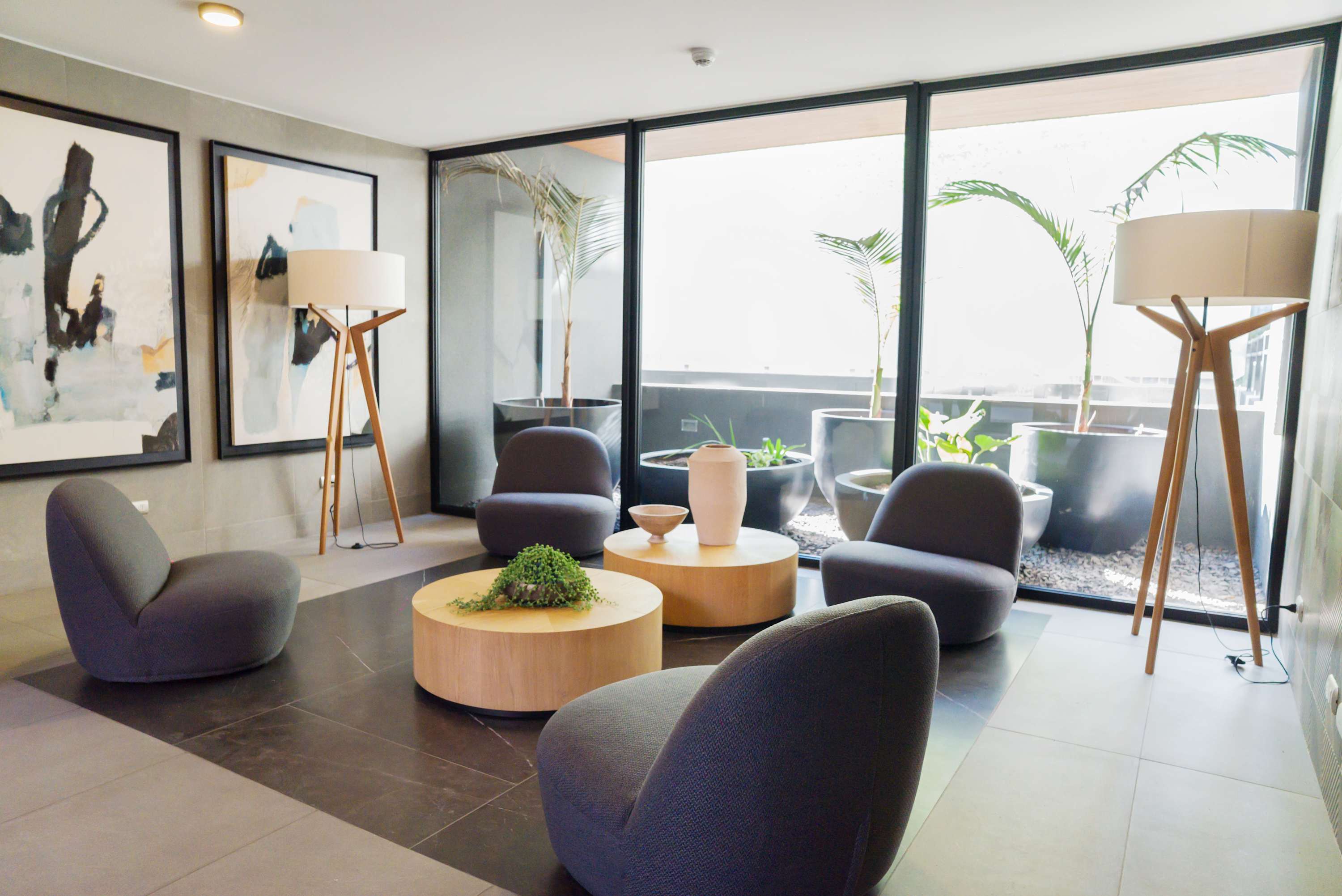 Contemporary lounge with sculptural seating, round wood tables, abstract artwork, and floor-to-ceiling glass overlooking a planted outdoor area.