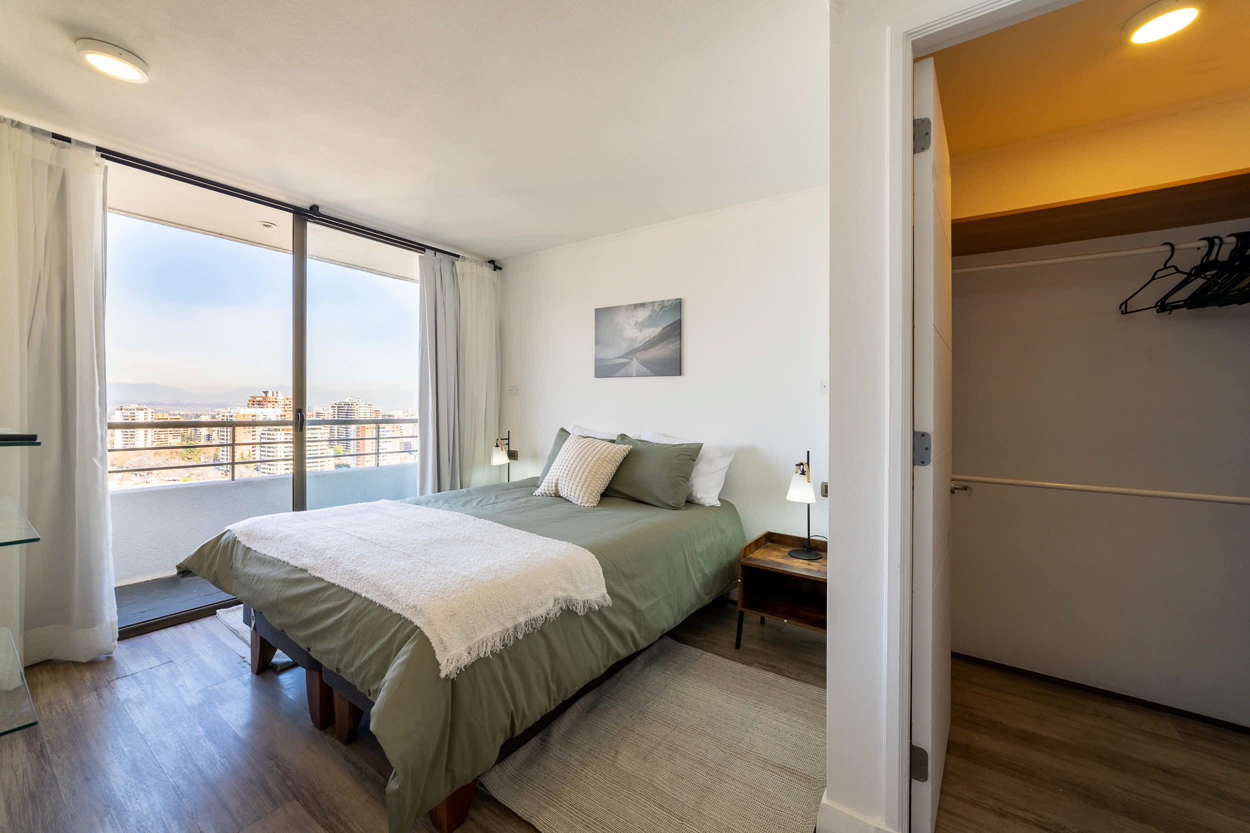 Bright bedroom with a comfortable double bed, soft sage-and-cream linens, and a balcony overlooking the city and mountains.