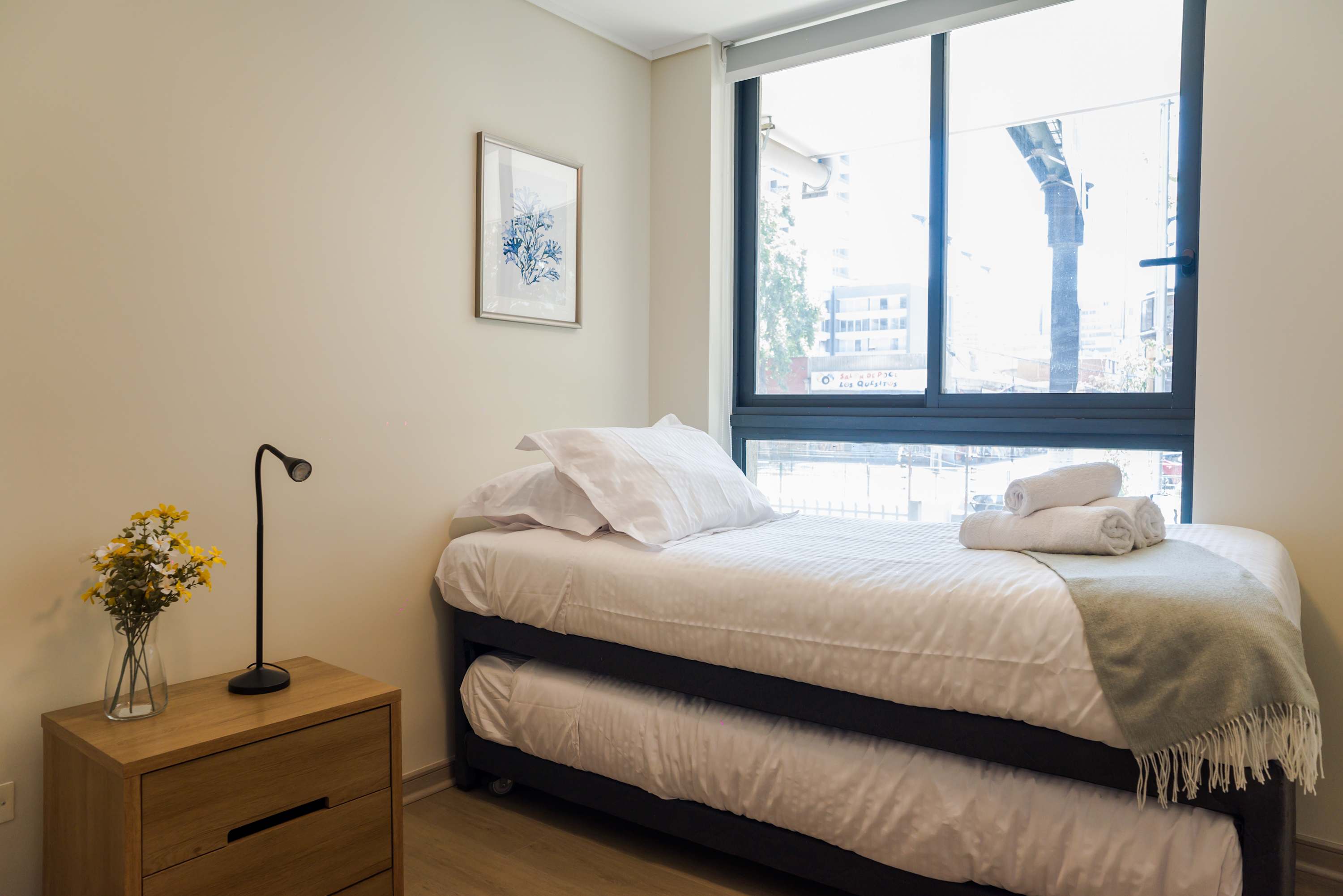 Private bedroom with a neatly made double bed, soft gray throw, fresh towels, and a large window overlooking the city.