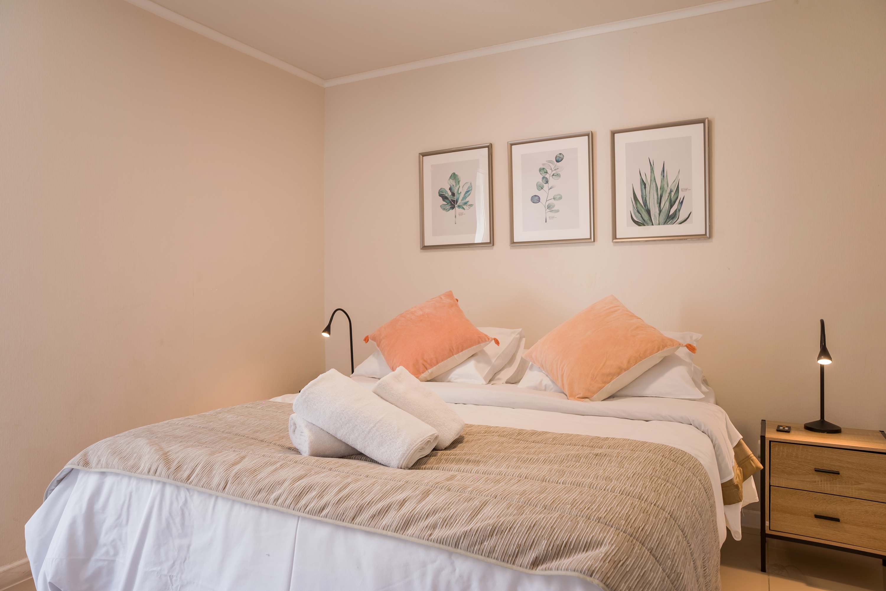 Private bedroom with crisp white bedding, coral accent pillows, folded towels, botanical artwork, and sleek bedside lighting.