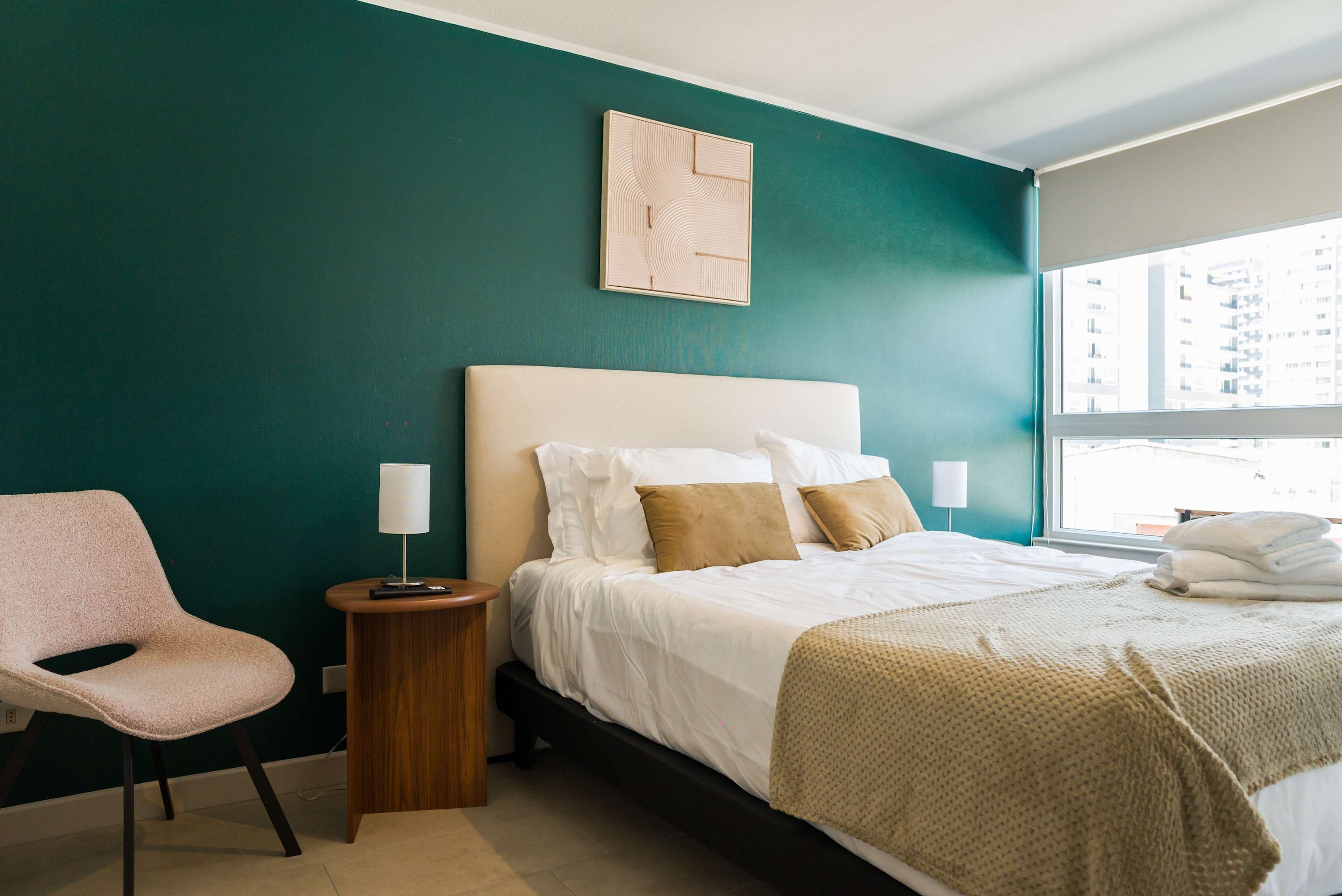 Private bedroom with a deep teal accent wall, layered white-and-tan bedding, and large windows overlooking the surrounding city buildings.