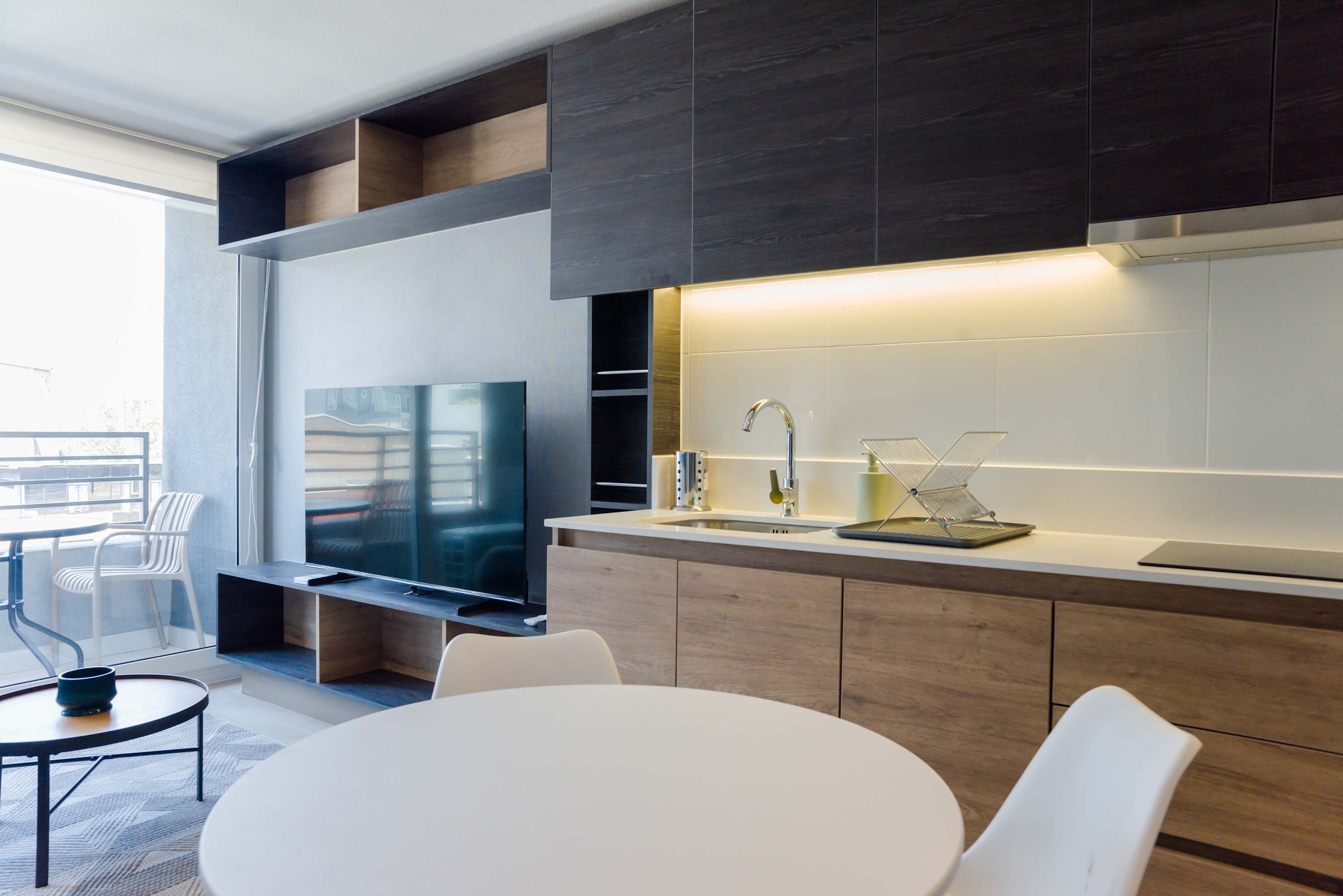 Modern kitchen and dining area with warm under-cabinet lighting, sleek wood cabinetry, and a sunlit balcony nook.