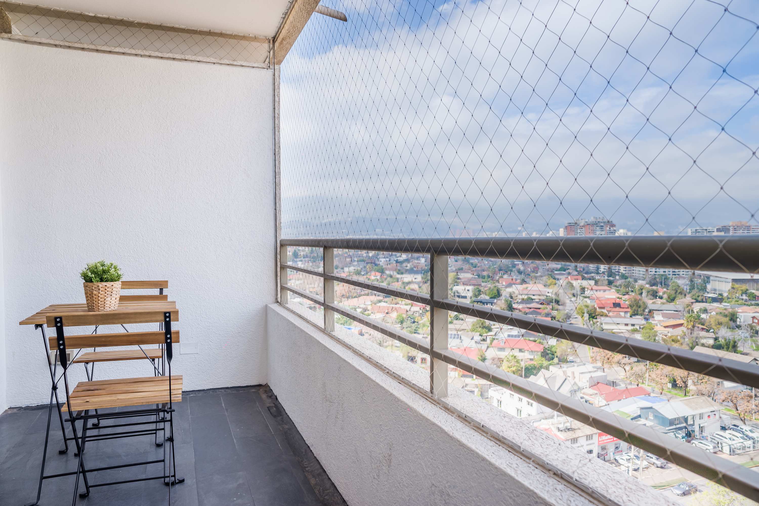 Private balcony with a two-seat wooden bistro set, potted greenery, and sweeping city views through protective safety netting.