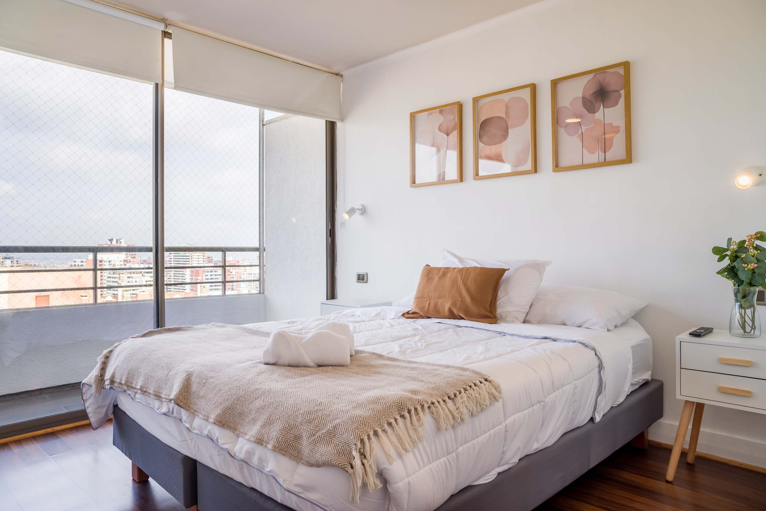 Bright bedroom with a large, comfortably dressed bed, floral artwork, and floor-to-ceiling windows overlooking the city.