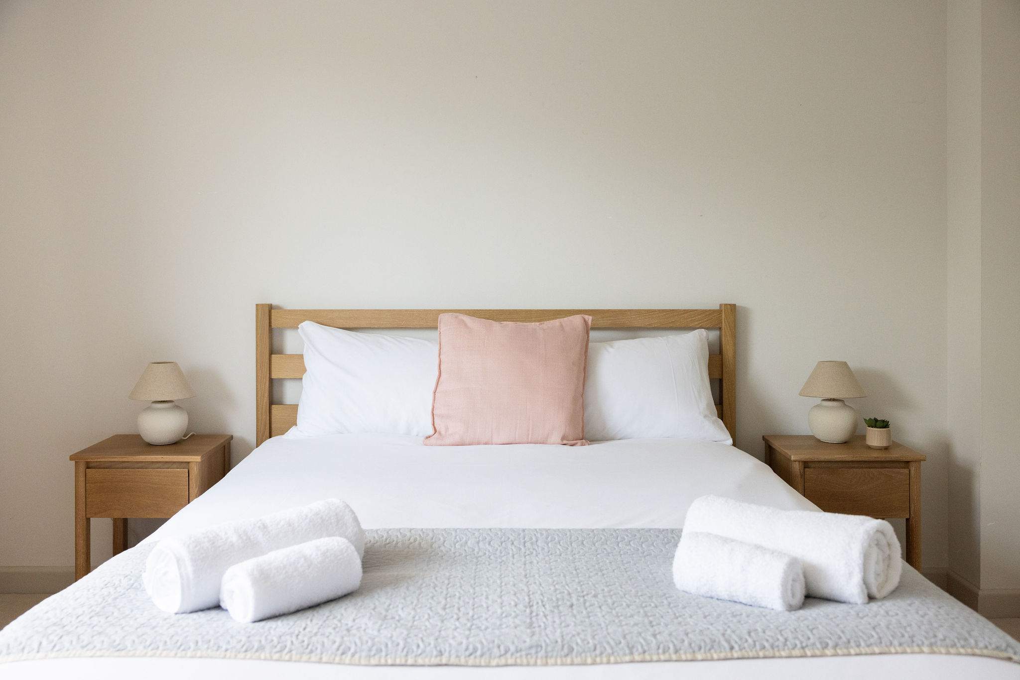 Bright, calming bedroom featuring a comfortable King bed, natural wood bedside tables, soft neutral bedding with a blush accent cushion, matching bedside lamps and fresh towels ready for your stay.