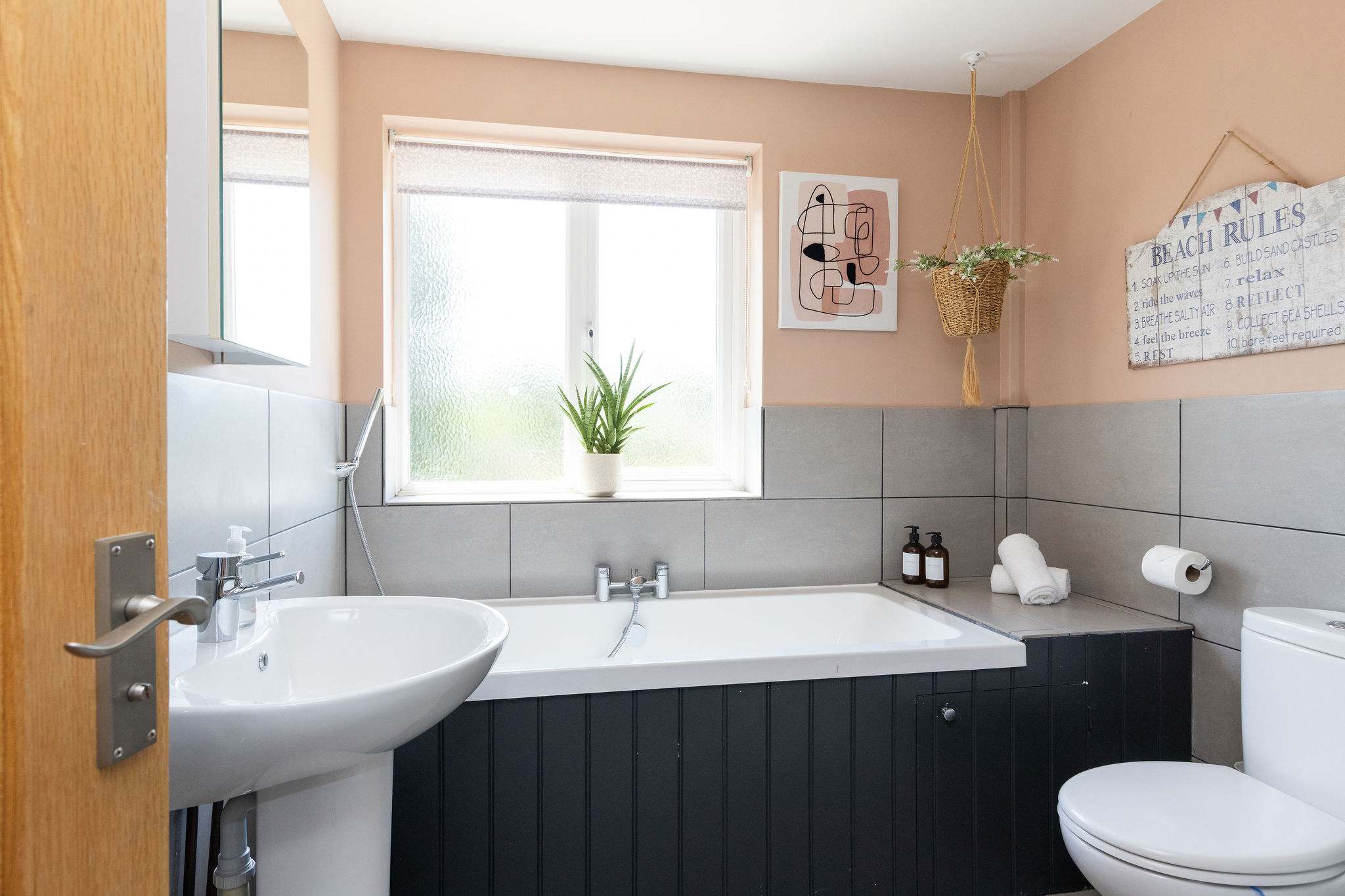 A fresh, well-appointed family bathroom with a full-size bath, sleek dark cabinetry, soft blush walls and contemporary grey tiling, all brought together with plenty of natural light.