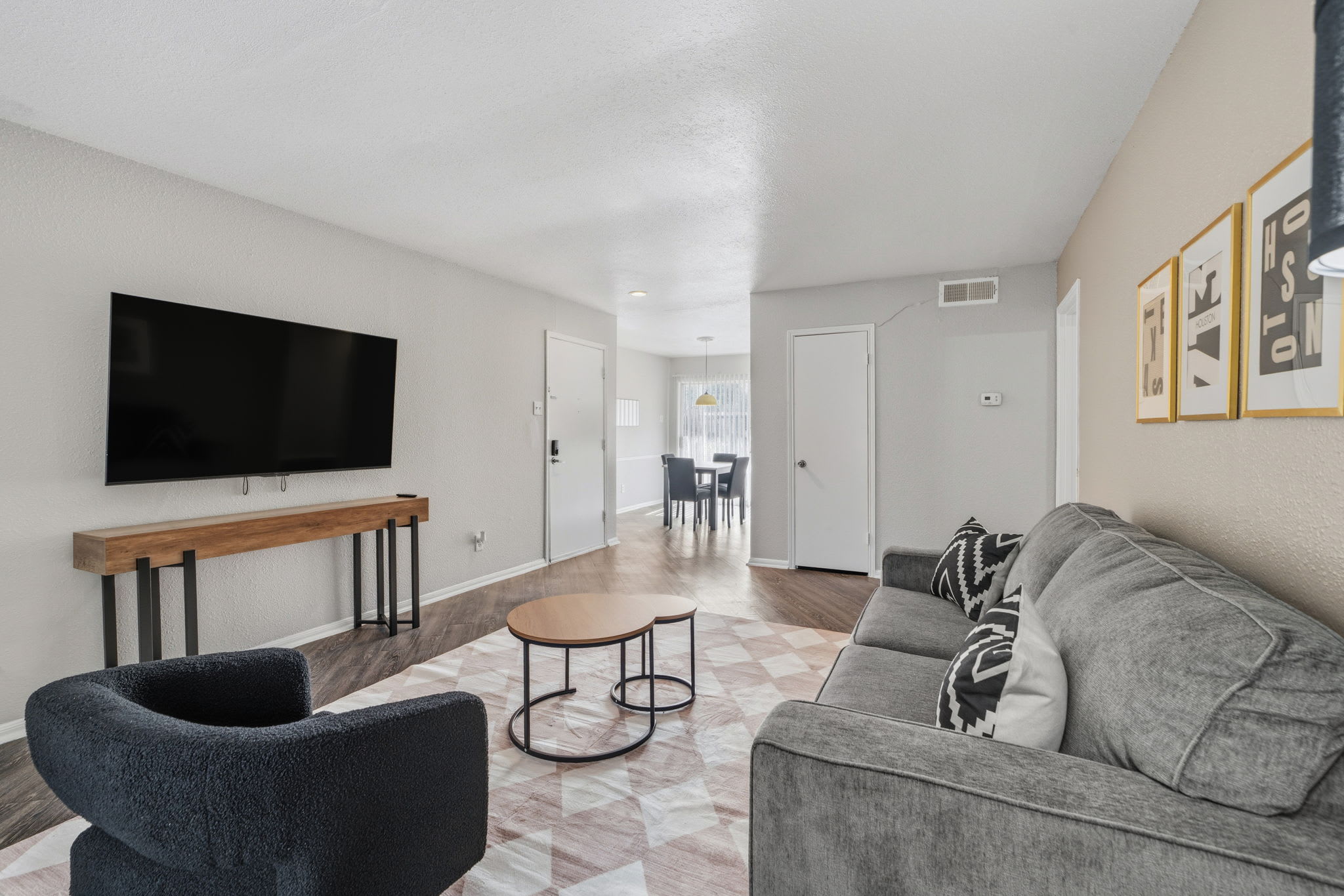 Private living room with a wall-mounted TV, gray sectional, geometric rug, and connected dining area filled with natural light.