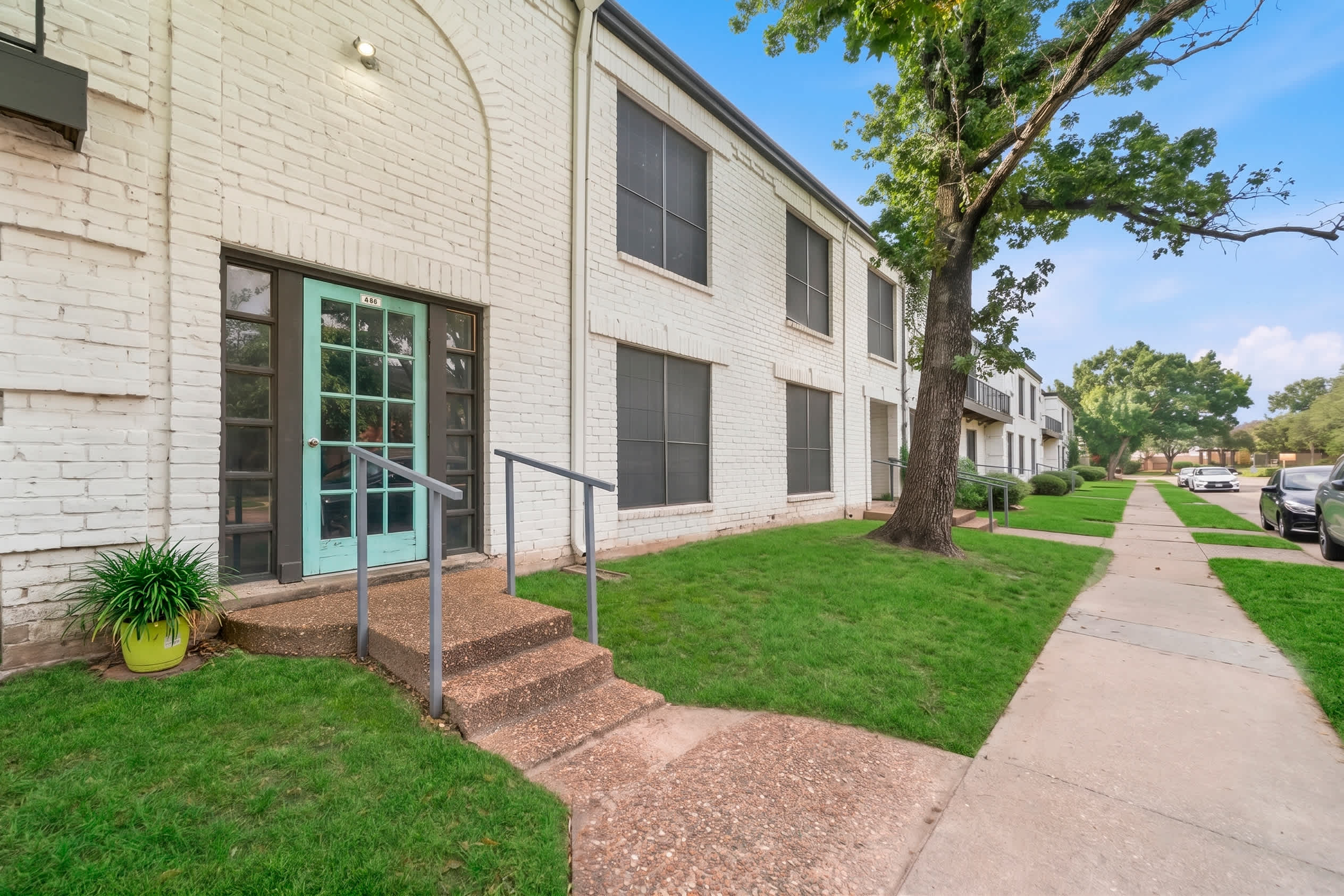 Welcoming apartment entrance with a turquoise glass-paneled door, landscaped lawn, mature shade tree, and accessible walkway.