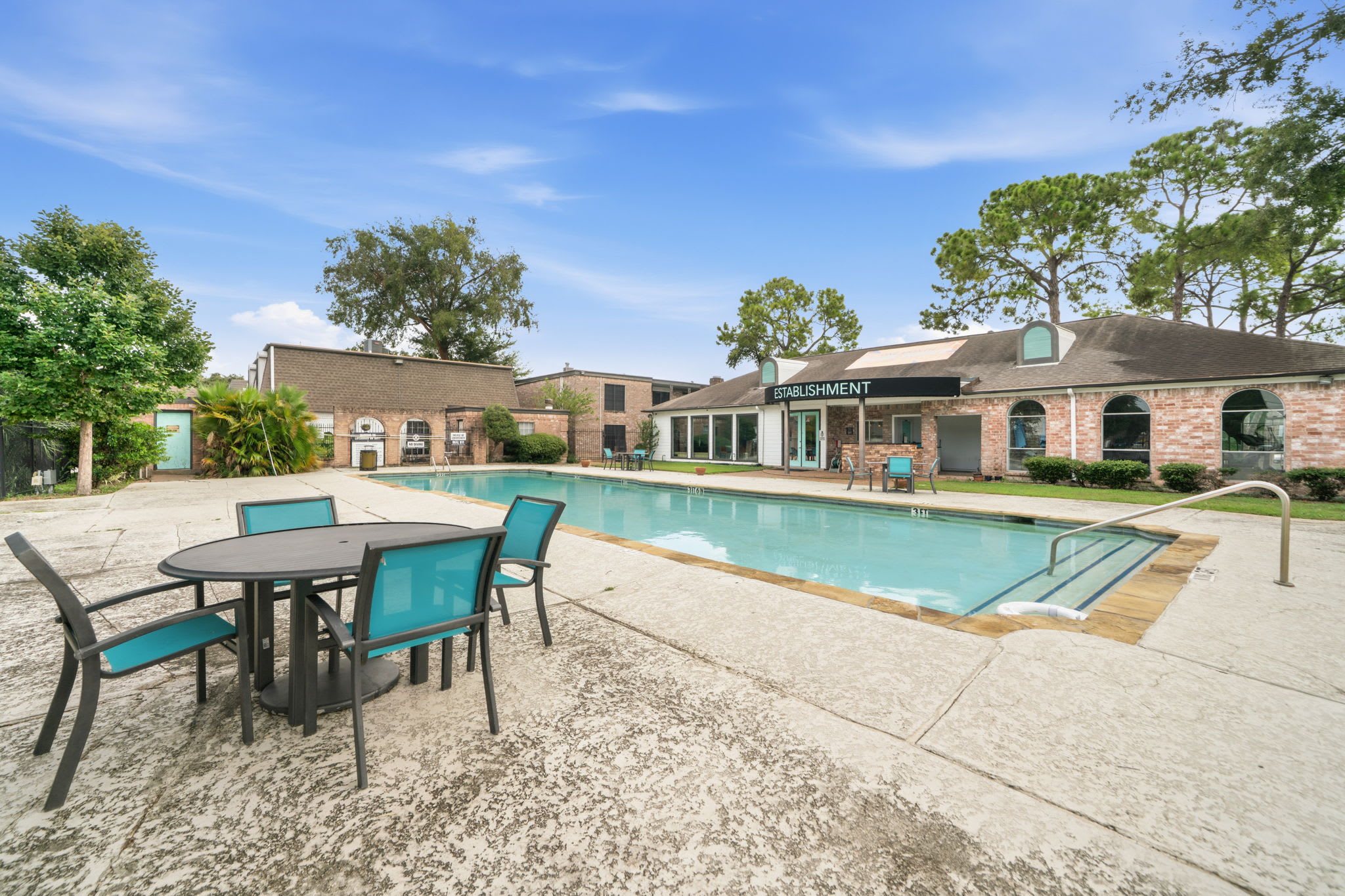 Shared pool with turquoise water, a shallow entry, and shaded outdoor dining surrounded by mature trees and brick buildings.