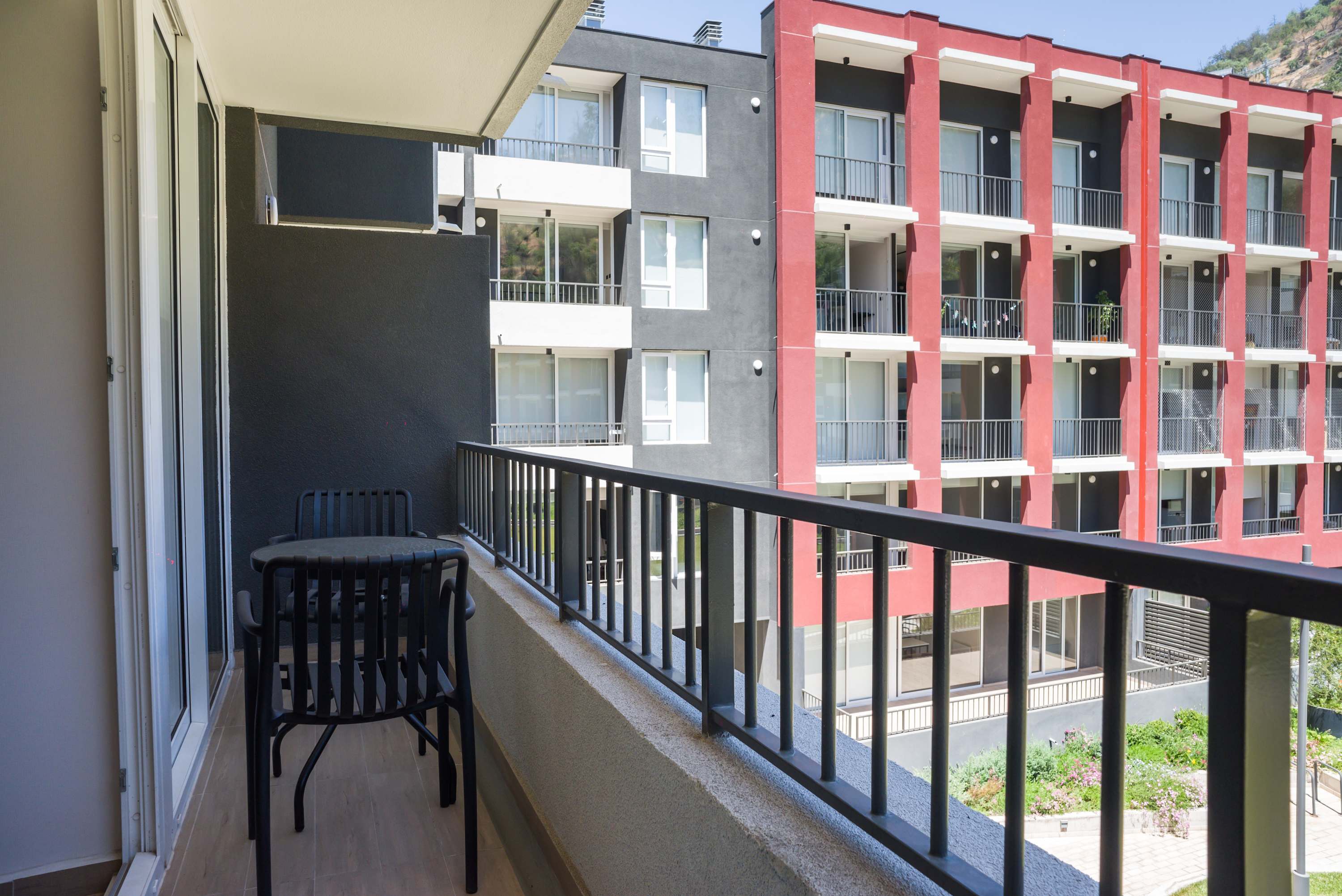 Sunny private balcony with a three-seat bistro set, overlooking landscaped grounds, modern apartments, and a wooded hillside.