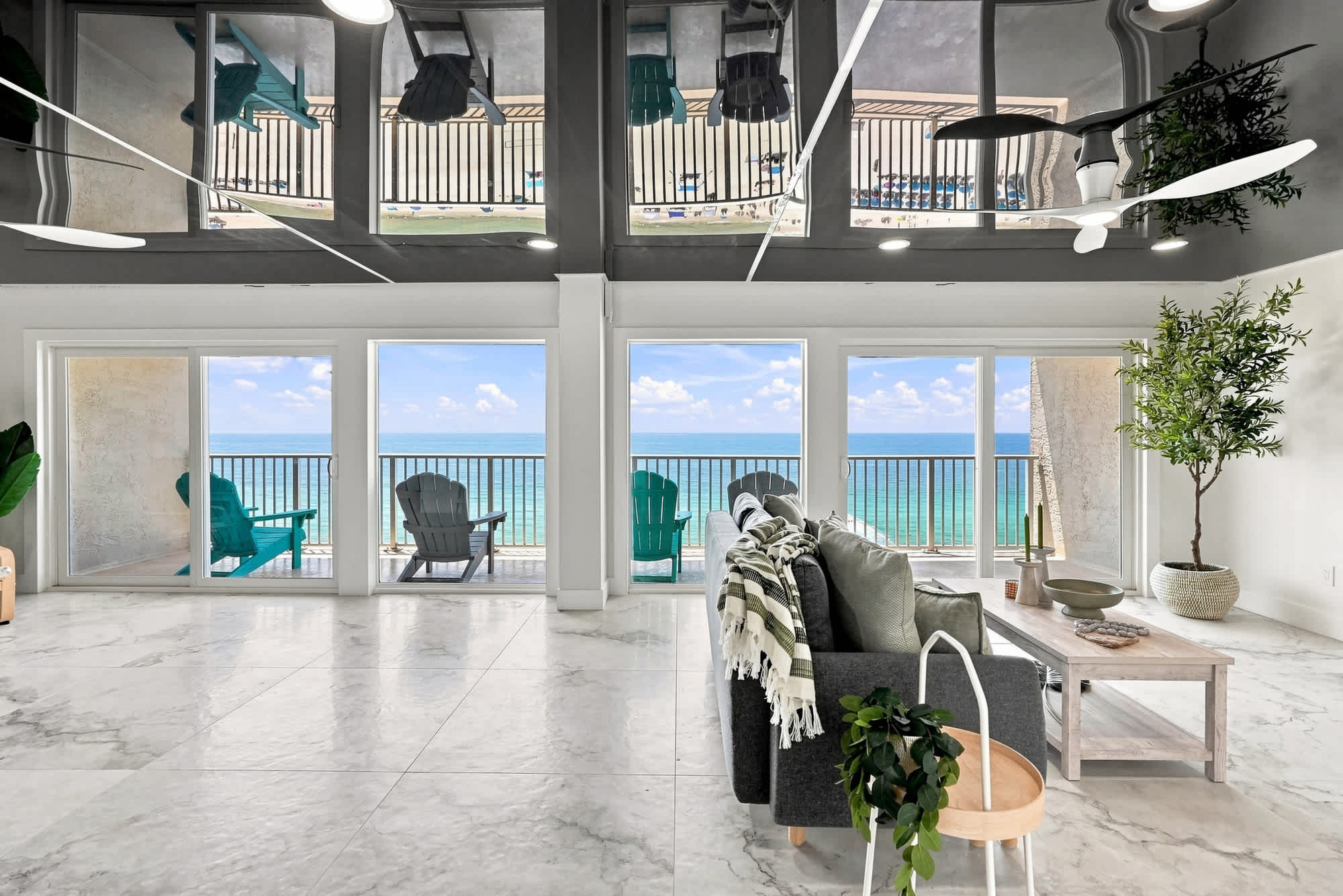 The Summit 1512 – Bright living room with ocean view, large windows, gray sofa, and teal chairs on the balcony.