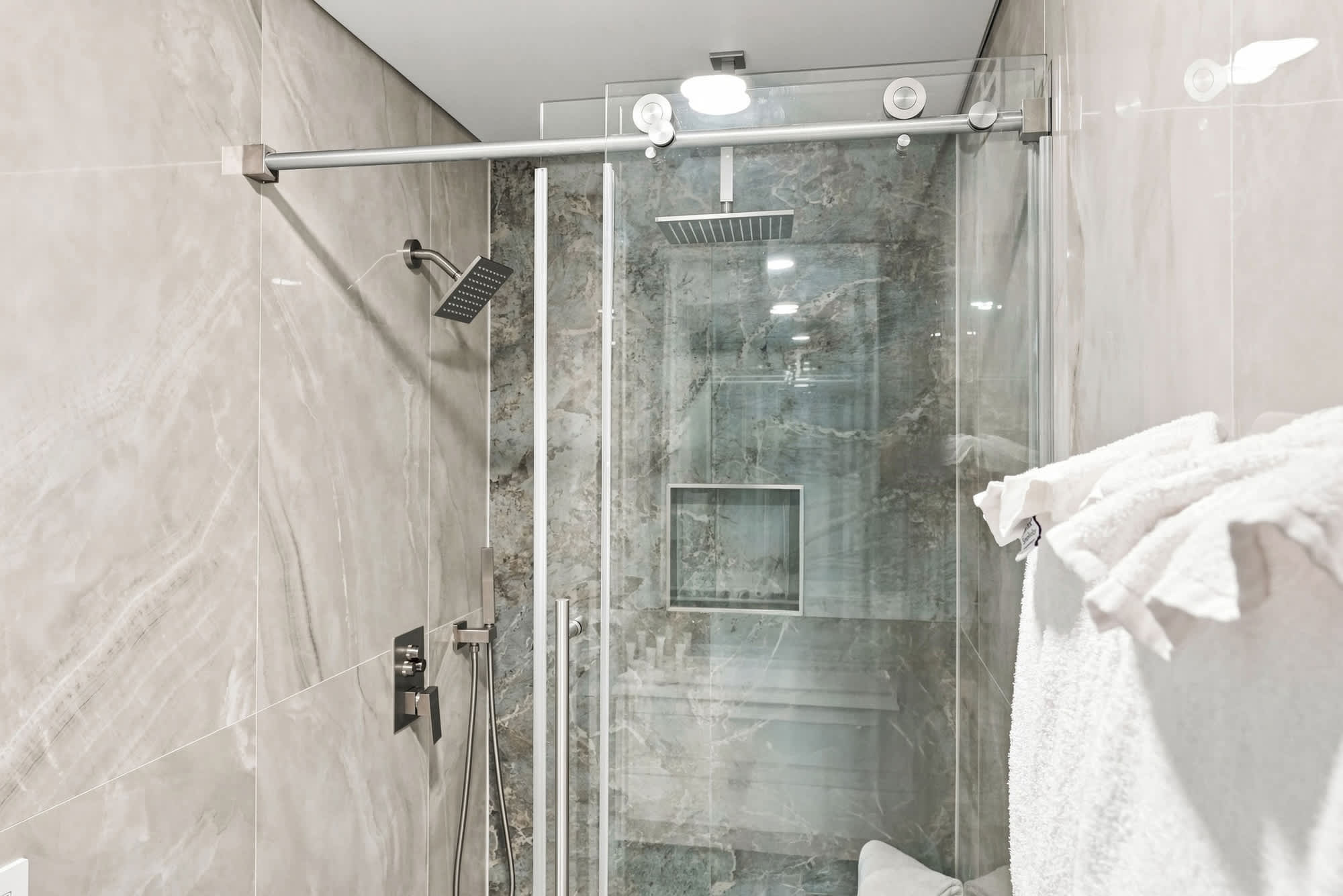The Summit 1512 – Modern glass shower with overhead and handheld fixtures, marble walls, and white towels.