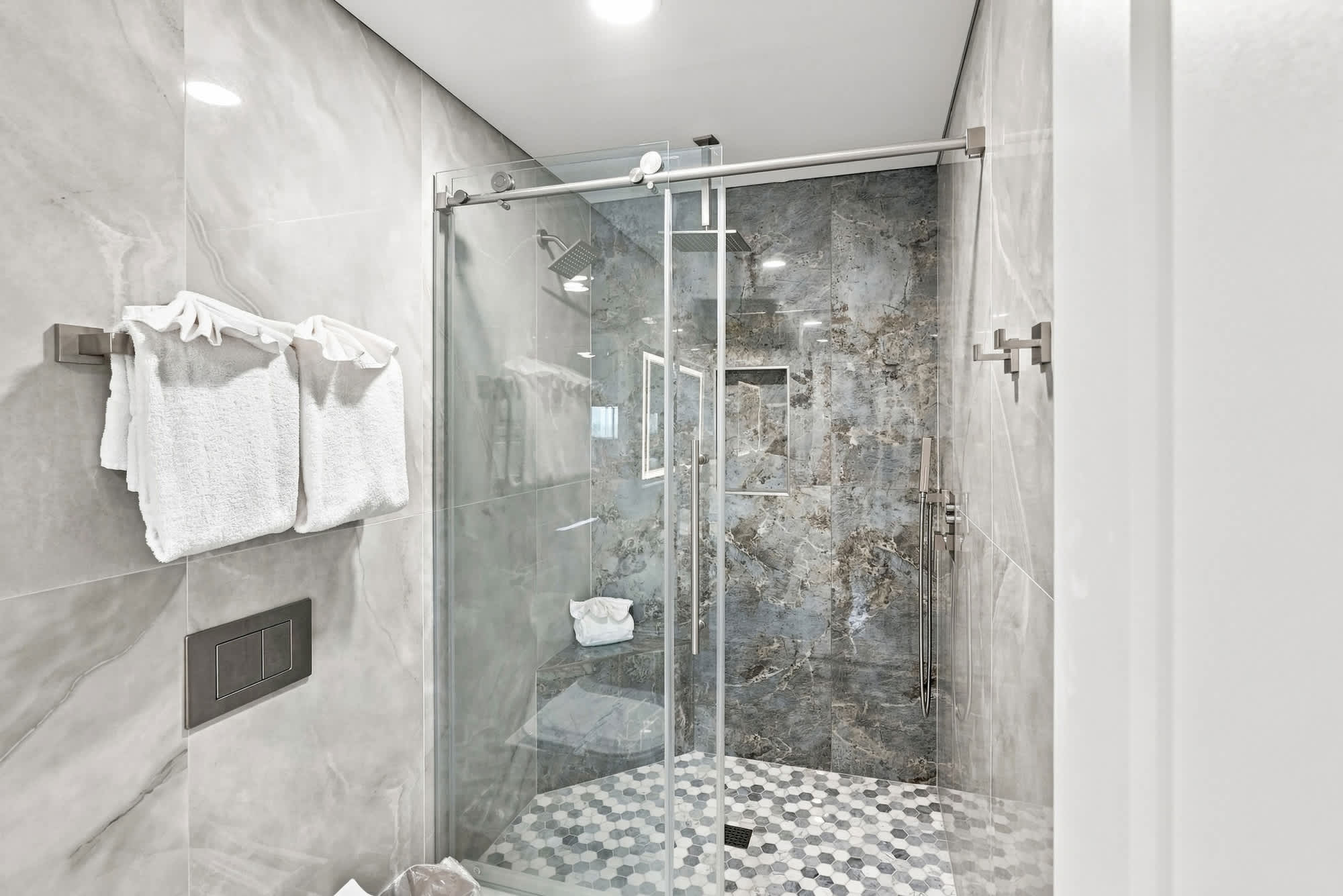The Summit 1512 – Modern bathroom with a glass shower, marble walls, hexagonal tile floor, and hanging towels.