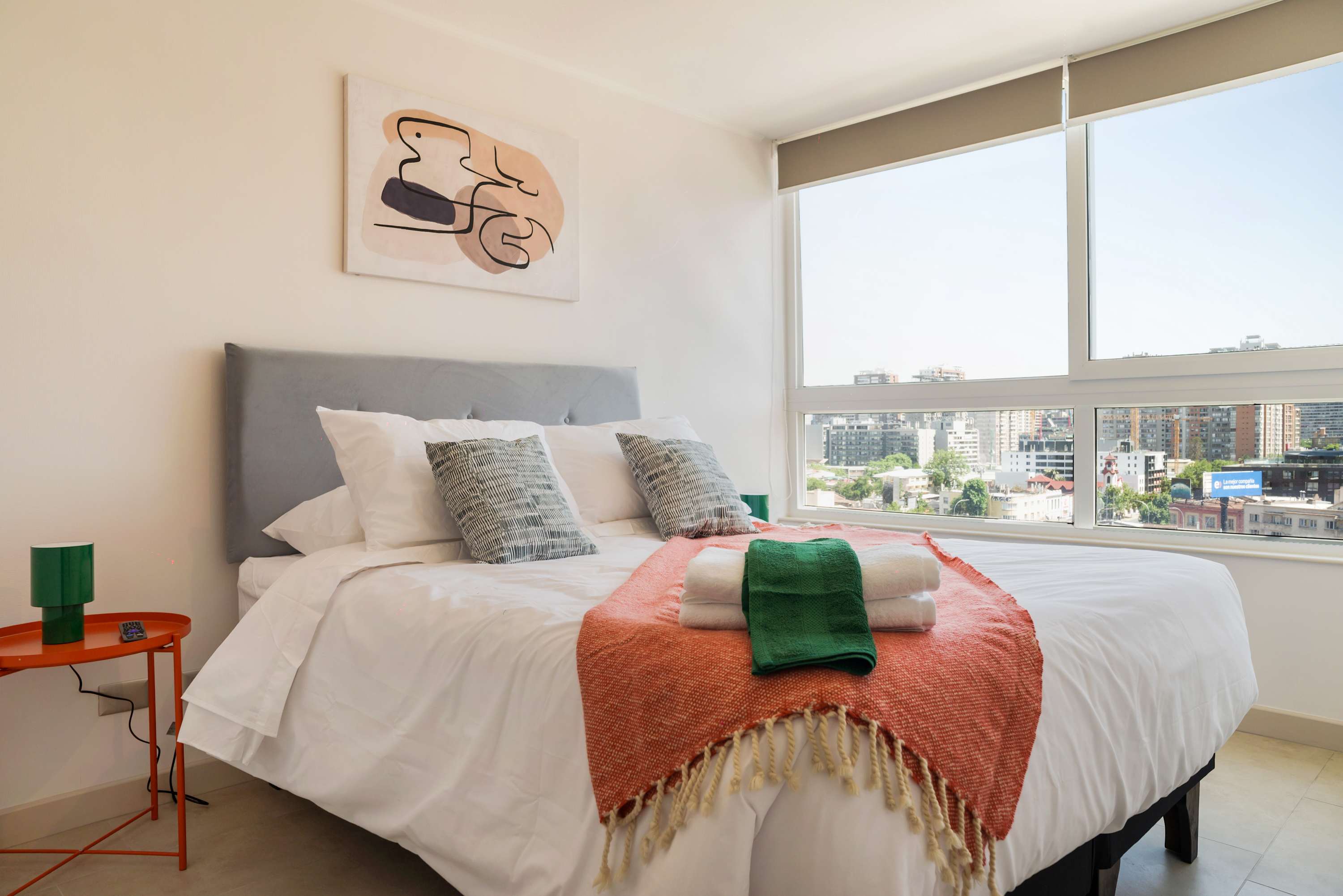 Private bedroom with crisp white bedding, colorful textured accents, and expansive city views through floor-to-ceiling corner windows.