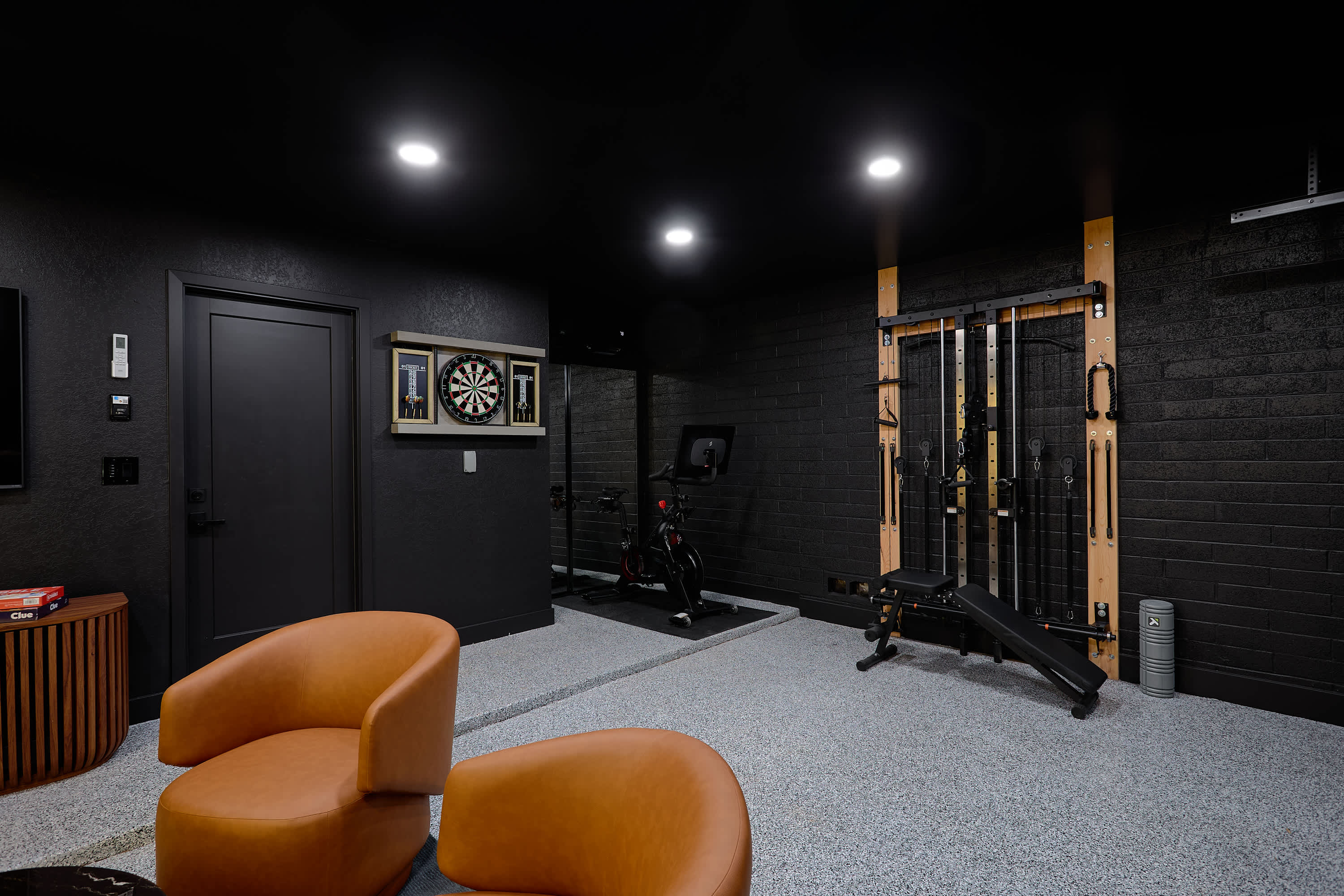 Game room and home gym packed with indoor entertainment, featuring a Peloton bike, ping pong table, arcade game, darts, TV, lounge seating, and a wall-mounted gym system.