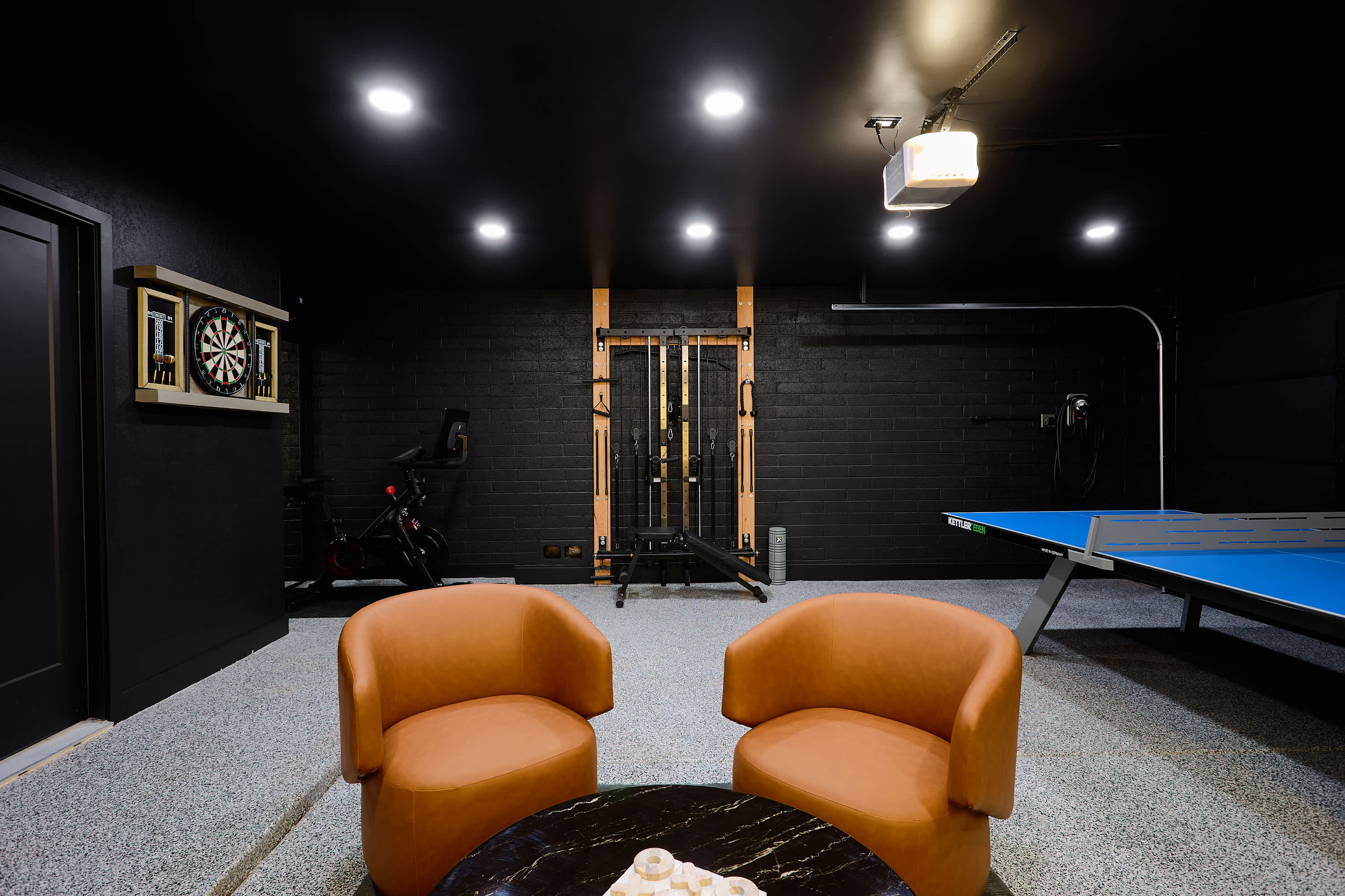 Game room and home gym packed with indoor entertainment, featuring a Peloton bike, ping pong table, arcade game, darts, TV, lounge seating, and a wall-mounted gym system.