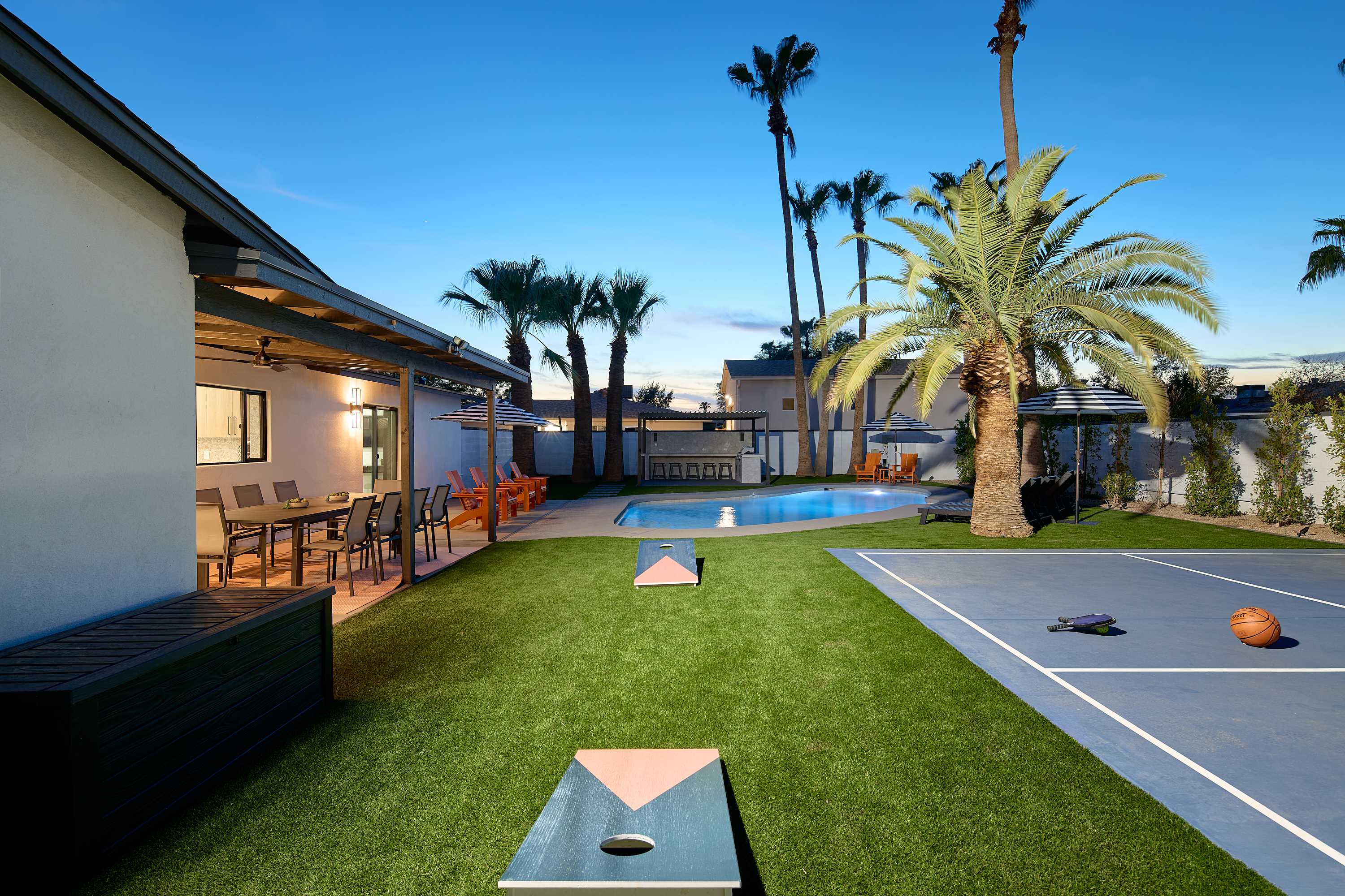 Resort-style backyard featuring a private pool, poolside loungers, a multi-sport court for basketball, pickleball, and tennis, turf with cornhole, a pergola with a built-in BBQ and bar seating, and a covered patio with outdoor dining table for 10.