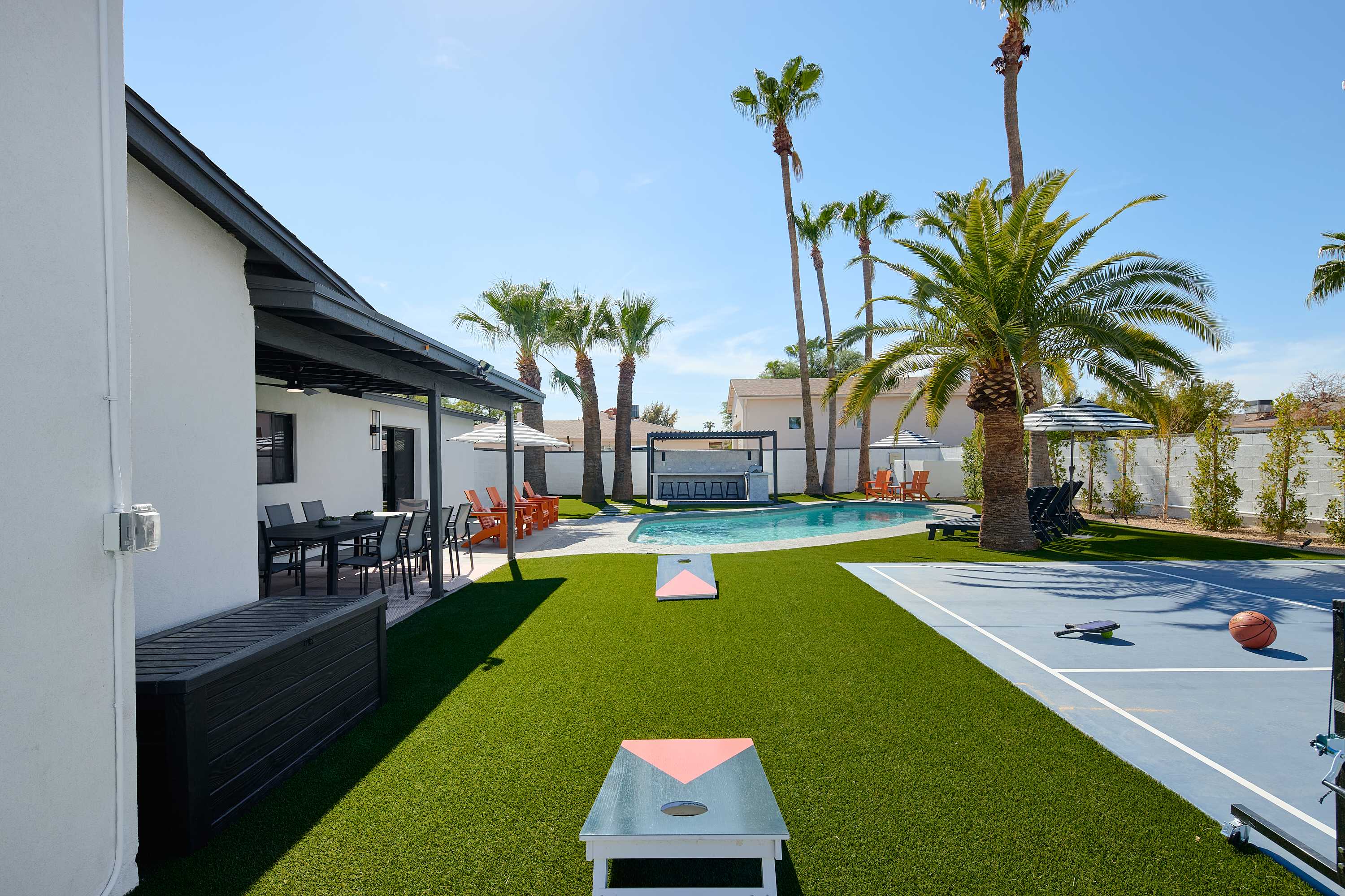 Resort-style backyard featuring a private pool, poolside loungers, a multi-sport court for basketball, pickleball, and tennis, turf with cornhole, a pergola with a built-in BBQ and bar seating, and a covered patio with outdoor dining table for 10.