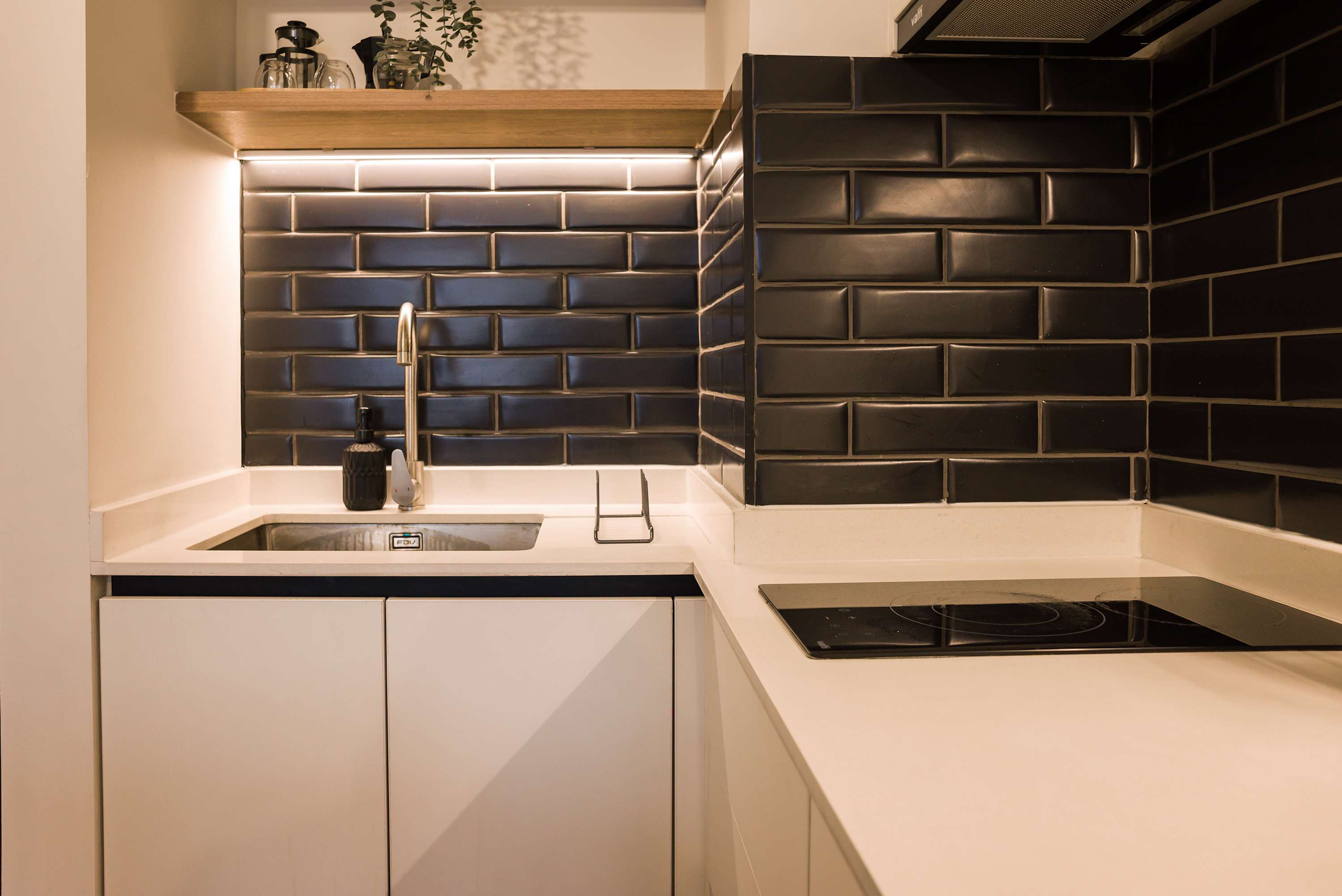 Compact private kitchen with a black induction cooktop, stainless-steel sink, glossy dark tile backsplash, and warm wood display shelf.