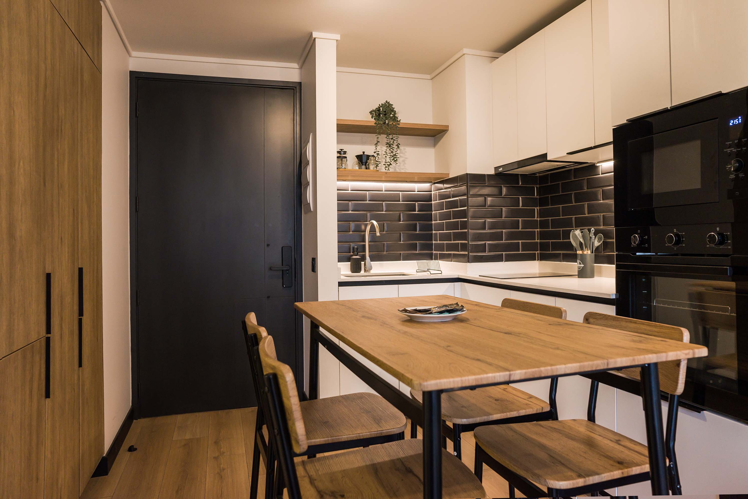 Private kitchen with a wood dining table, sleek black subway-tile backsplash, integrated appliances, and open shelves stocked with coffee and glassware.