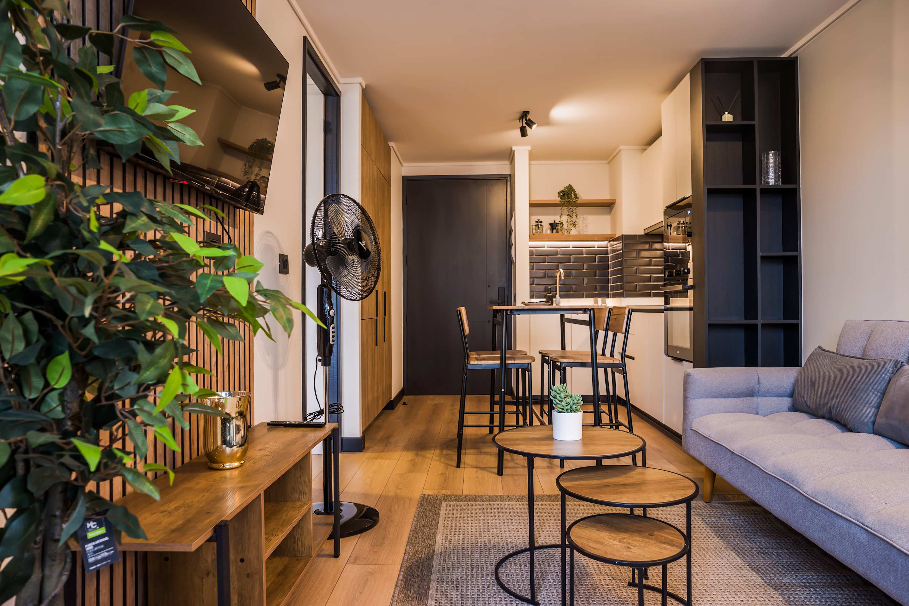 Open-plan living room with a gray sofa, nested wood coffee tables, and a stylish kitchen-dining area with four bar stools.