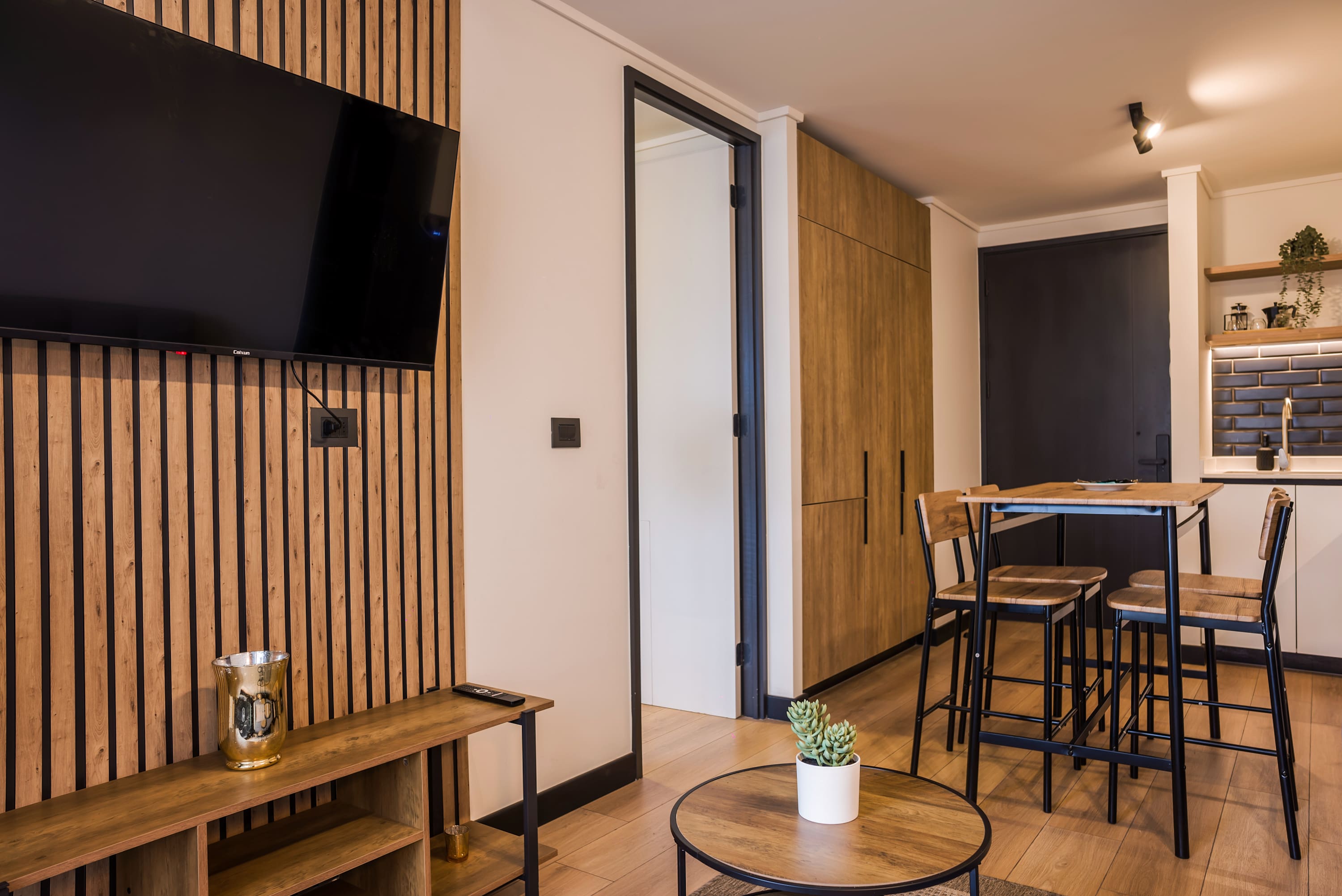 Open-plan living room with a large TV on a wood-slat accent wall, flowing into a modern kitchen and dining area.