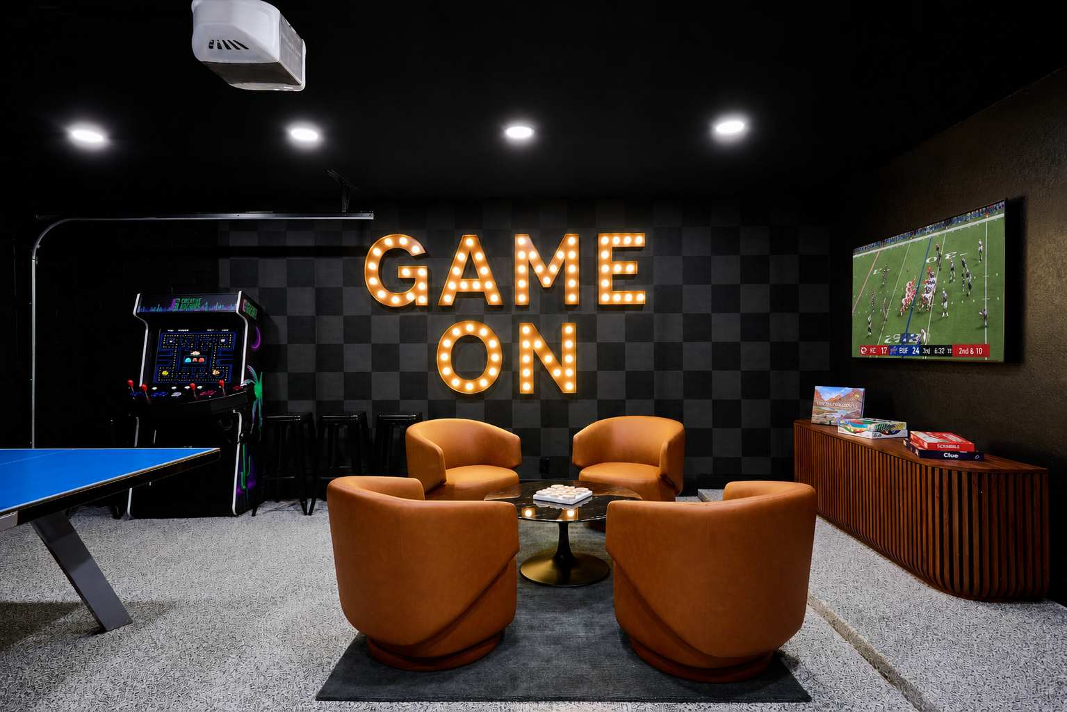 Game room and home gym packed with indoor entertainment, featuring a Peloton bike, ping pong table, arcade game, darts, TV, lounge seating, and a wall-mounted gym system.