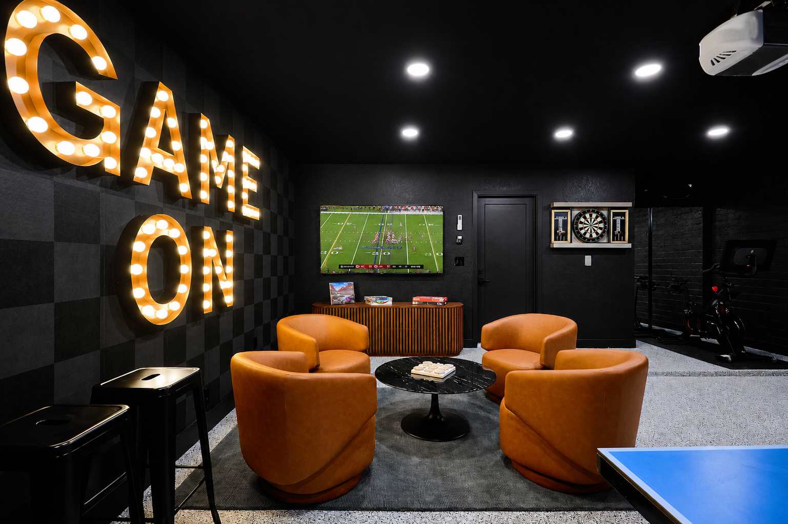 Game room and home gym packed with indoor entertainment, featuring a Peloton bike, ping pong table, arcade game, darts, TV, lounge seating, and a wall-mounted gym system.