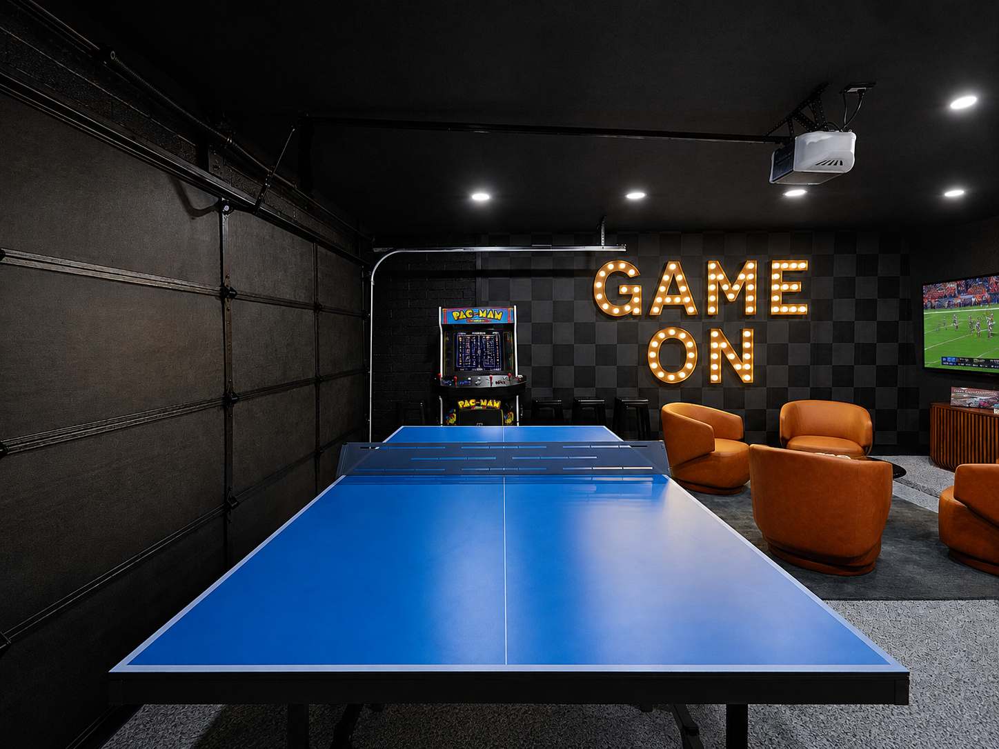 Game room and home gym packed with indoor entertainment, featuring a Peloton bike, ping pong table, arcade game, darts, TV, lounge seating, and a wall-mounted gym system.