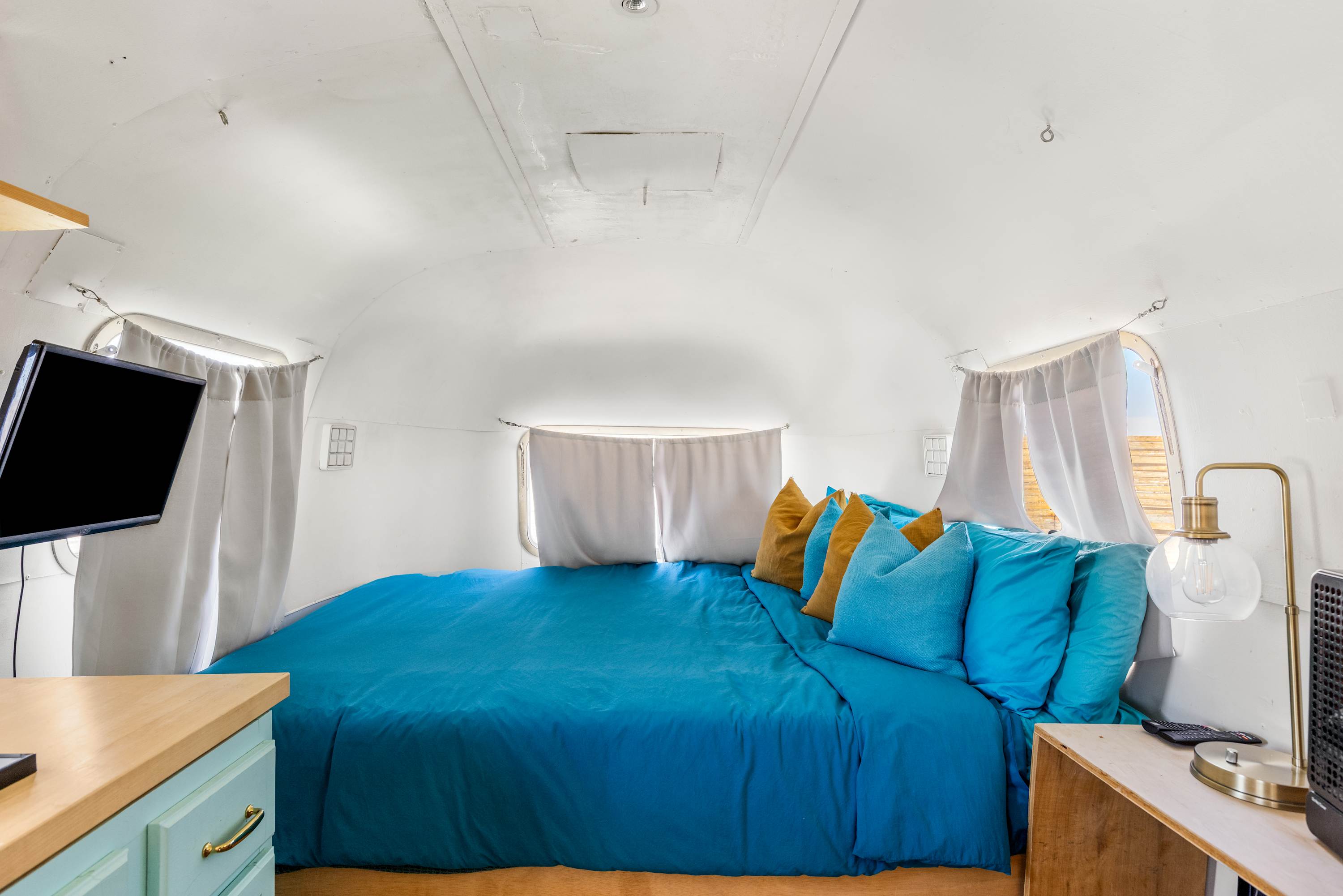 Airstream sleeping area with blue bedding, curtains, a bedside lamp and wall-mounted TV.