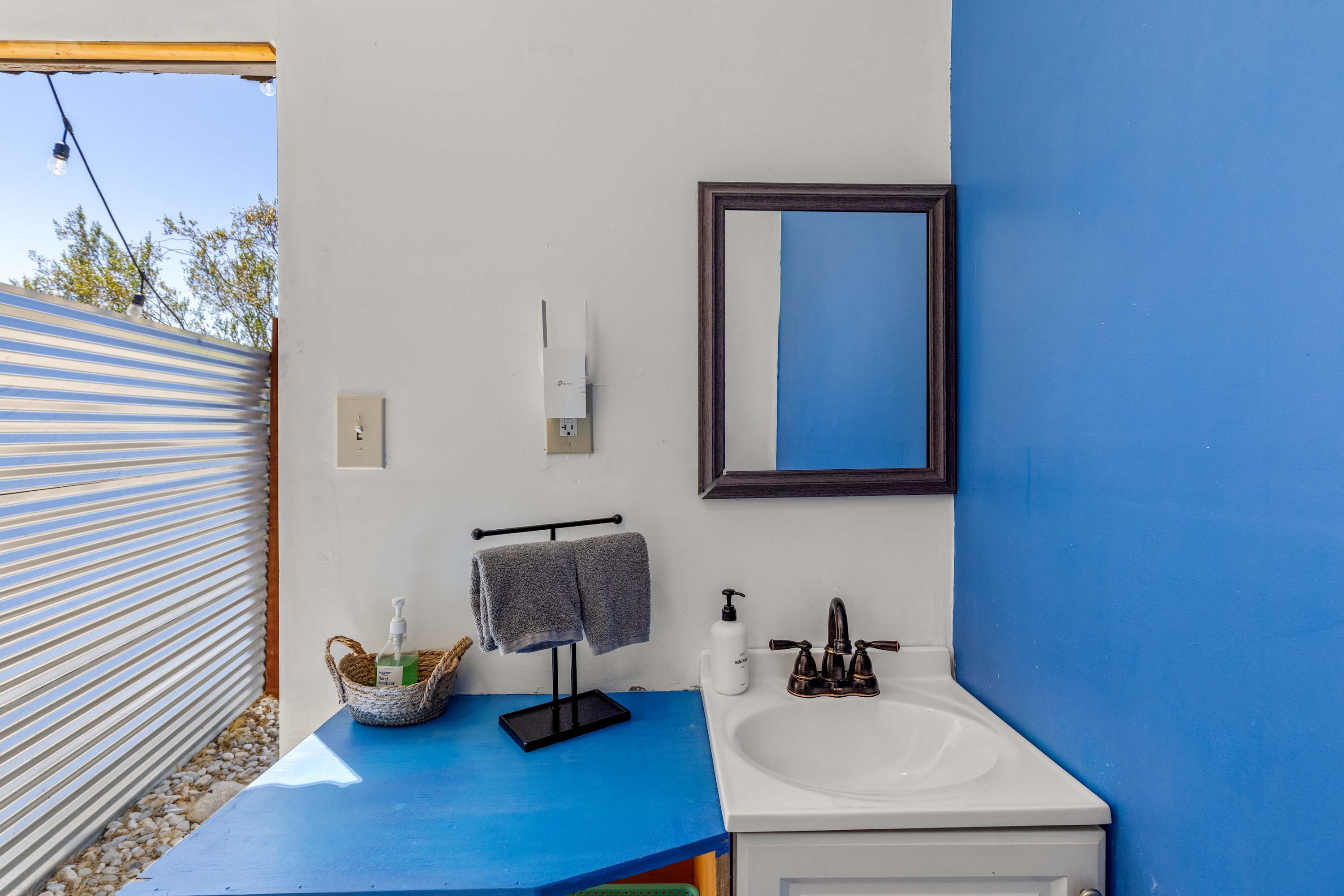 Outdoor bathroom vanity with a white sink, blue counter, mirror and towel rack.