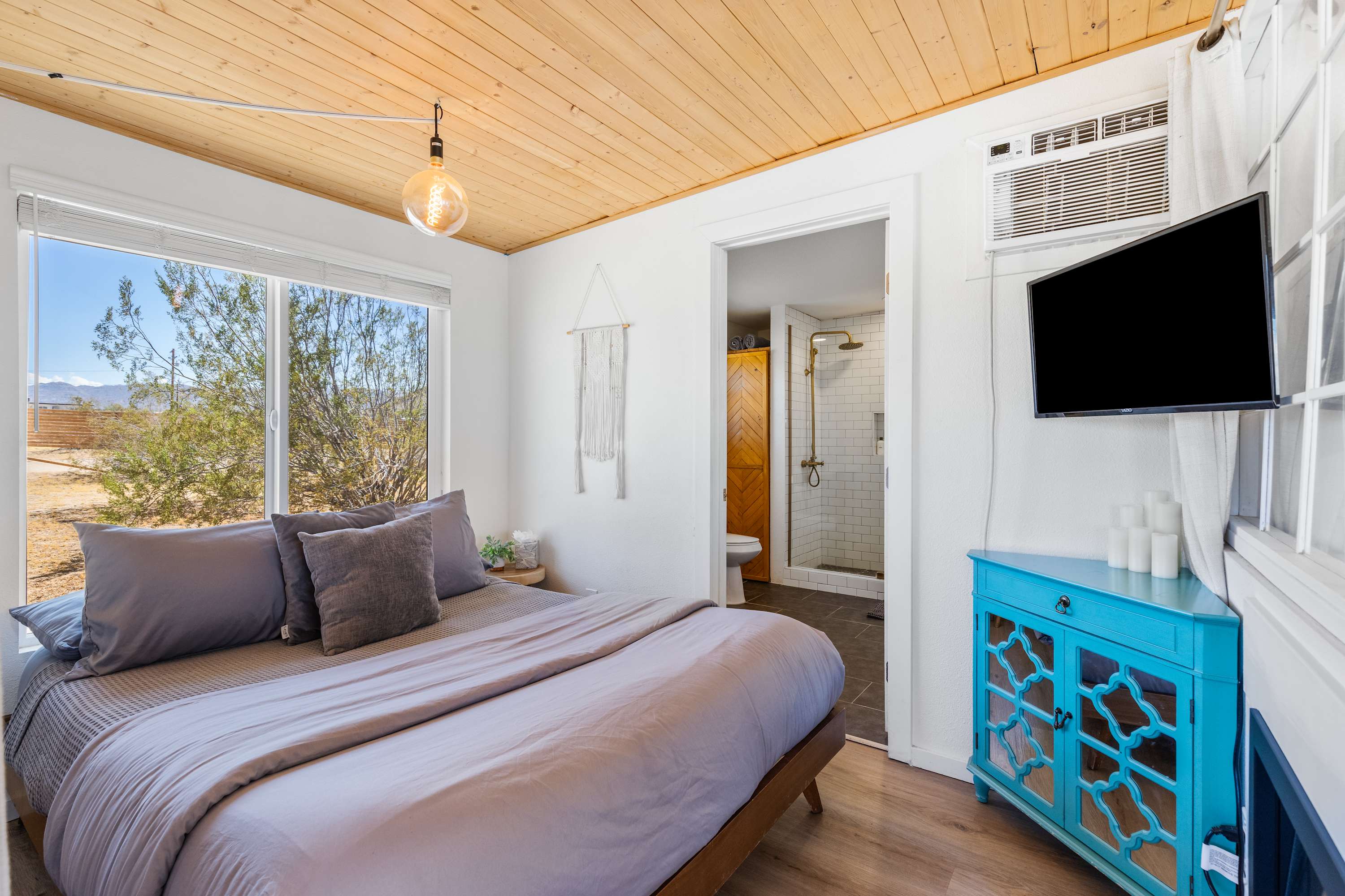 Studio sleeping area with a gray upholstered bed, wood ceiling, windows and a wall-mounted TV.