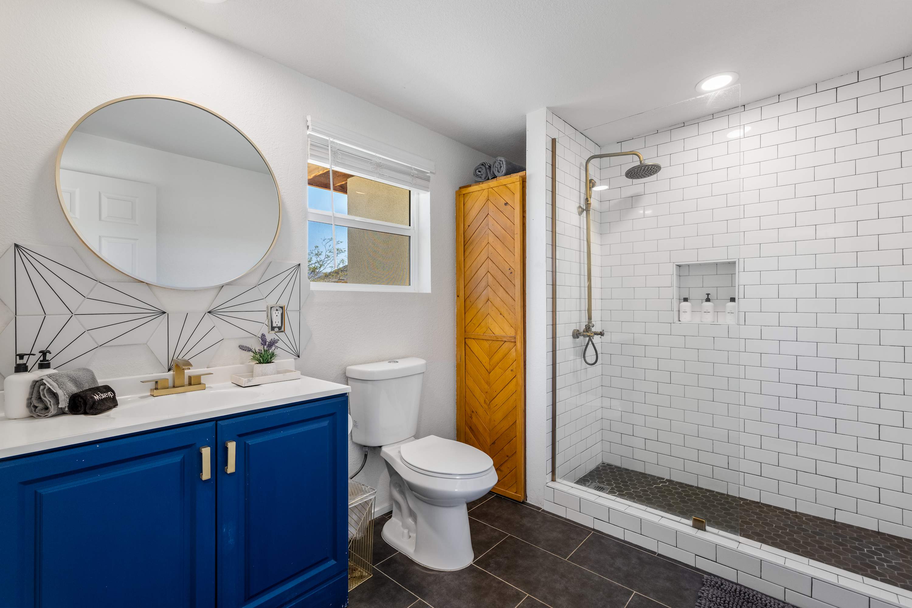 Studio bathroom with a navy vanity, round mirror, toilet and tiled walk-in shower.