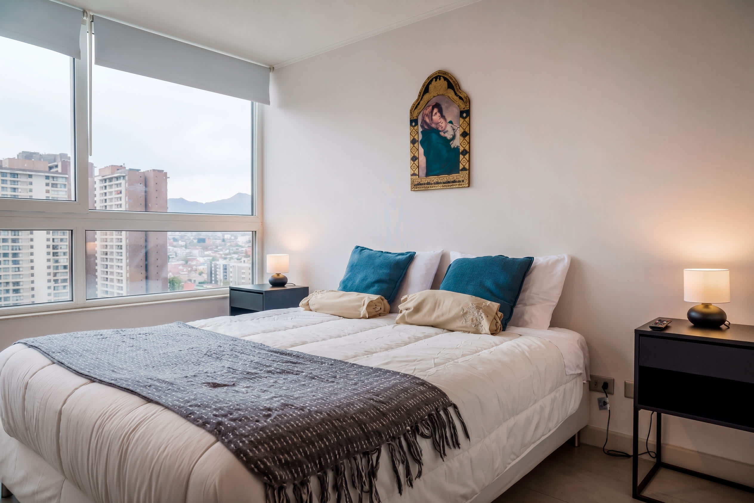 Bright bedroom with a neatly layered bed, teal accent pillows, warm bedside lighting, and expansive city-and-mountain views through the corner windows.