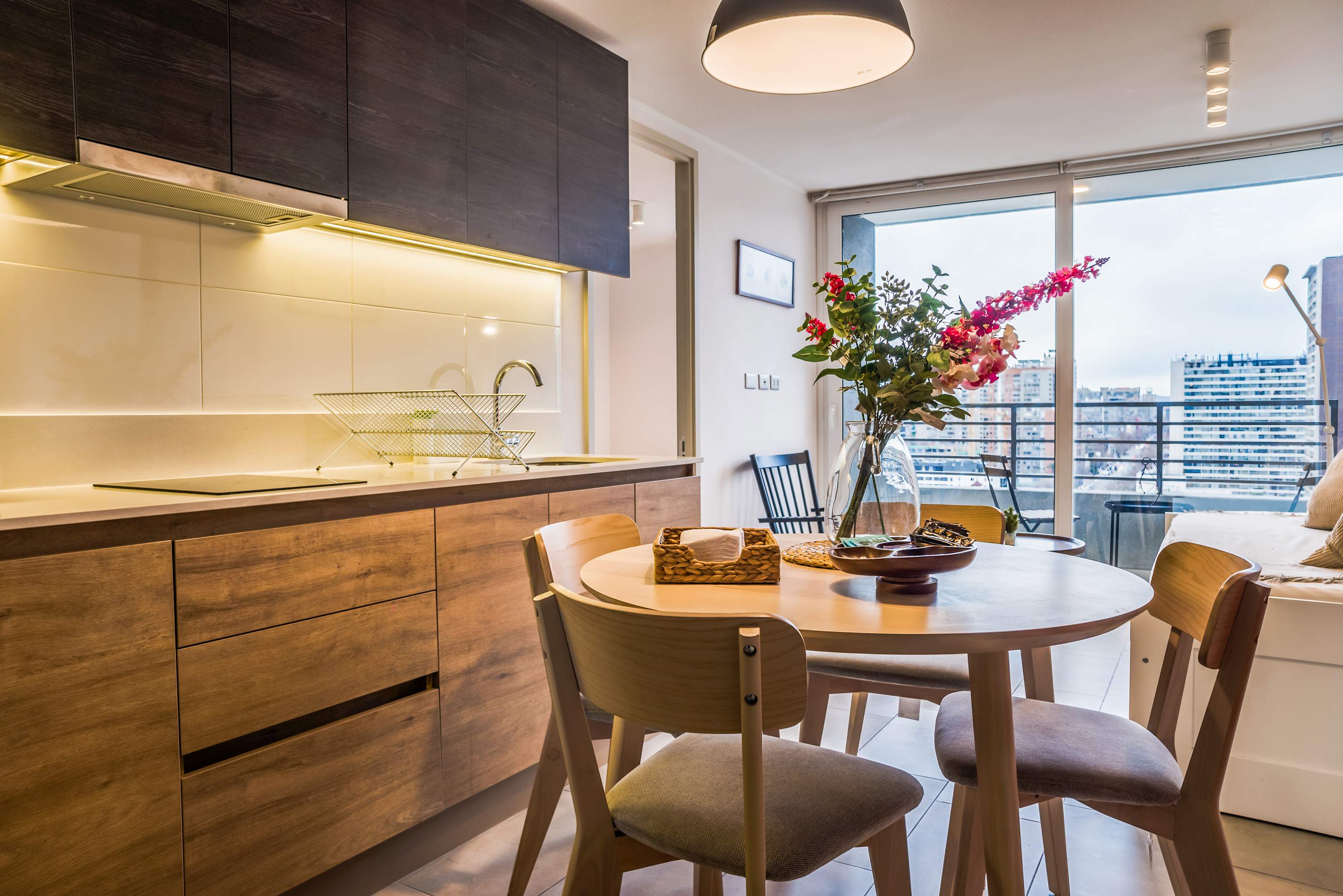 Modern kitchen and dining area with warm wood cabinetry, a four-seat table, and bright city views through the balcony windows.