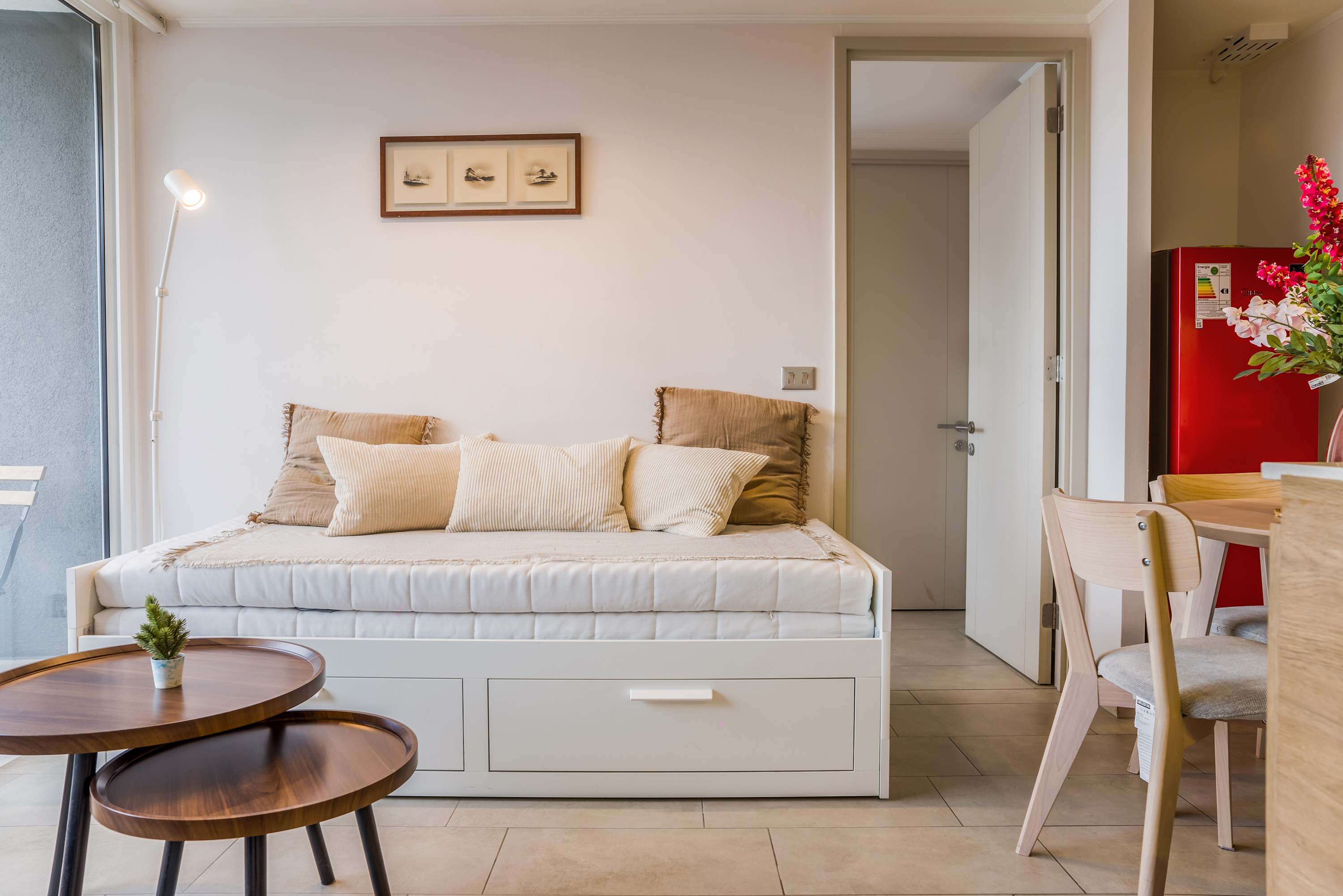 Bright living area with a comfortable white daybed, layered neutral cushions, round wood coffee tables, and a dining nook beside the vibrant red refrigerator.