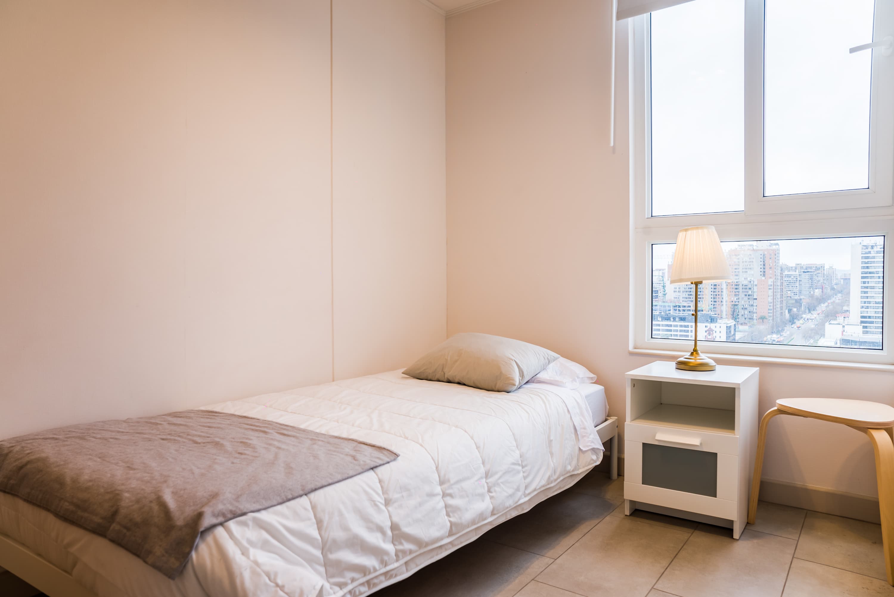 Bright private bedroom with a neatly made double bed, bedside lamp, and city views through large windows.