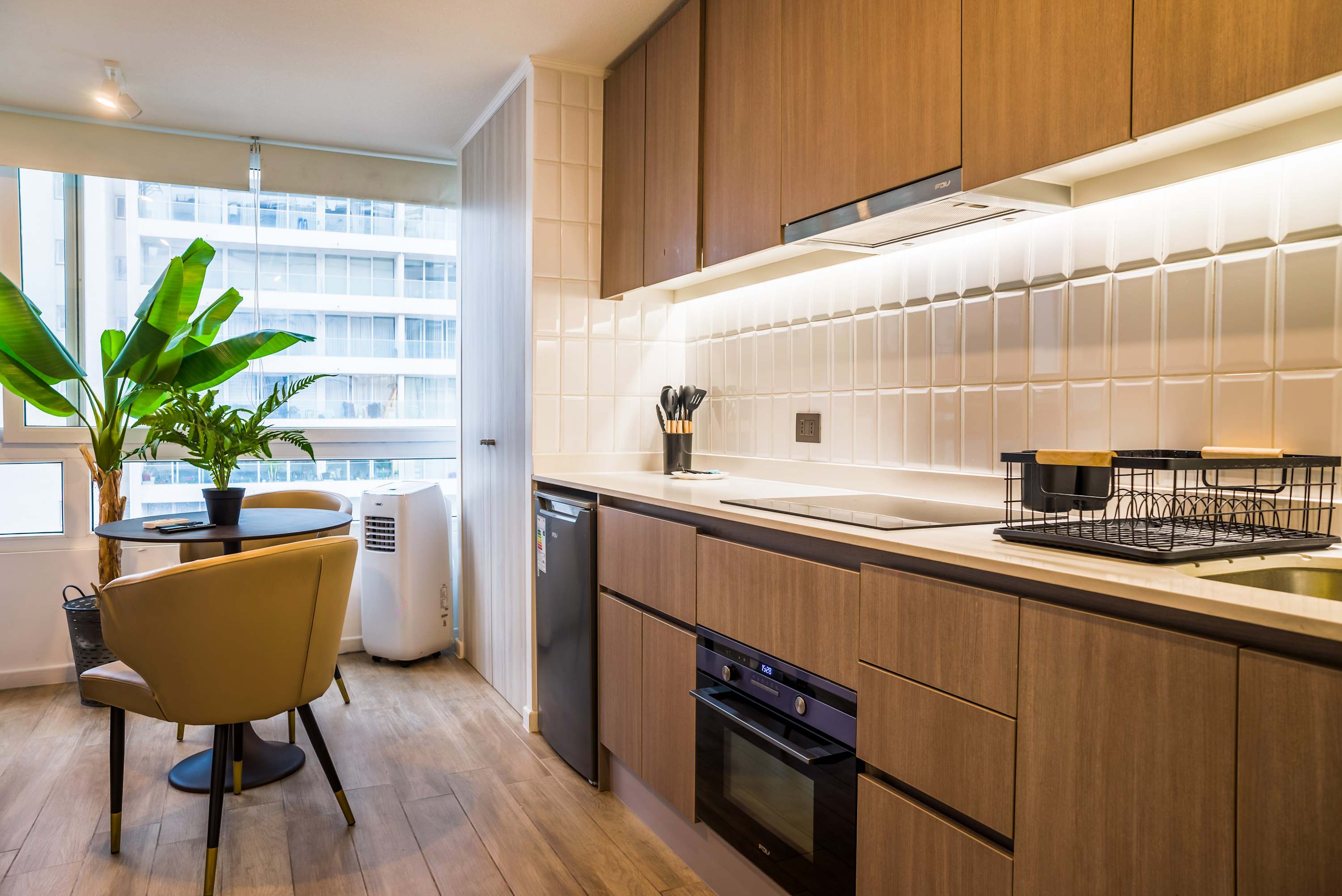 Bright studio kitchen with a two-seat dining table, modern wood cabinetry, glossy tile backsplash, and leafy plants beside the window.