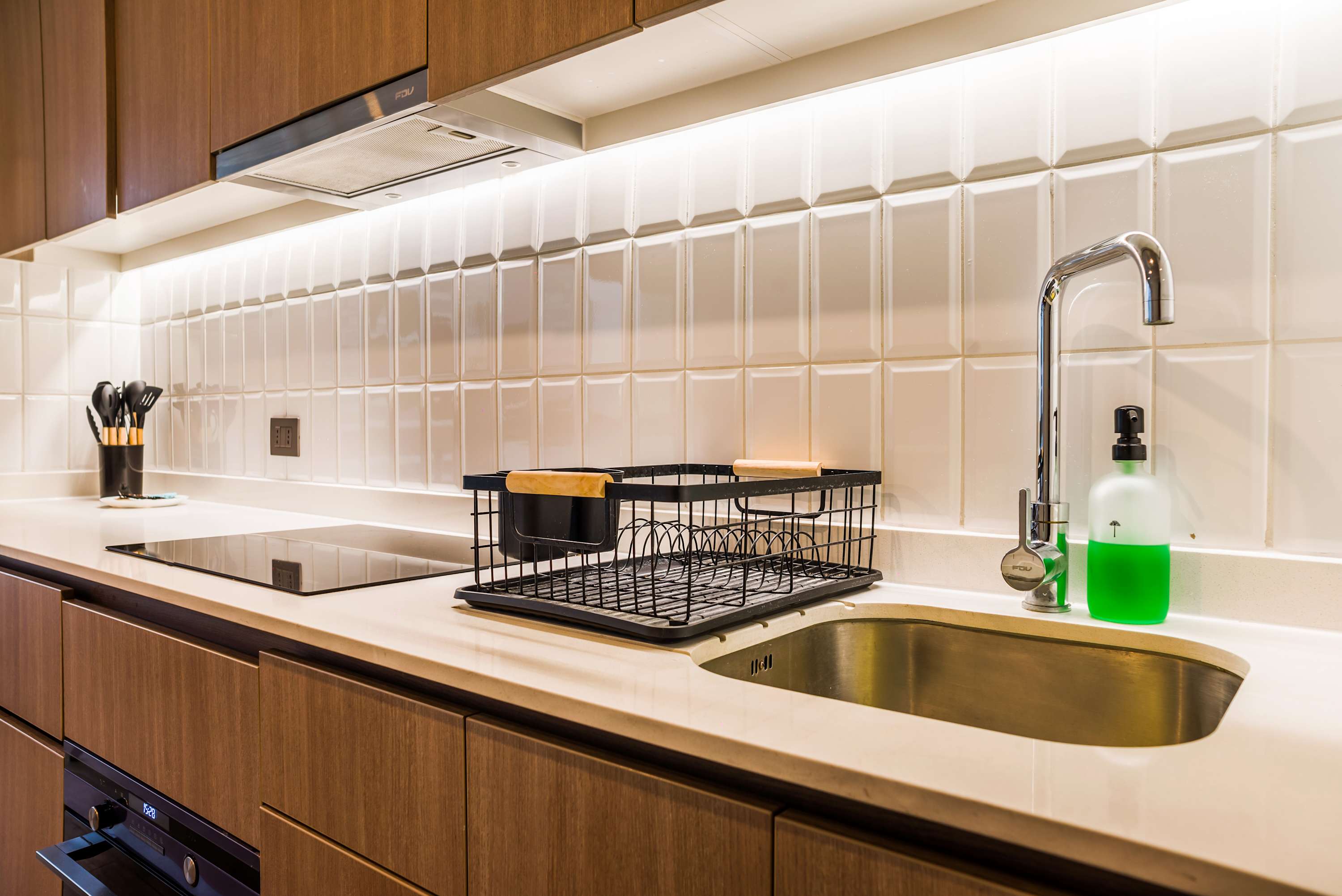 Compact studio kitchen with warm wood cabinetry, glossy white backsplash tiles, a built-in cooktop, and stainless-steel sink.