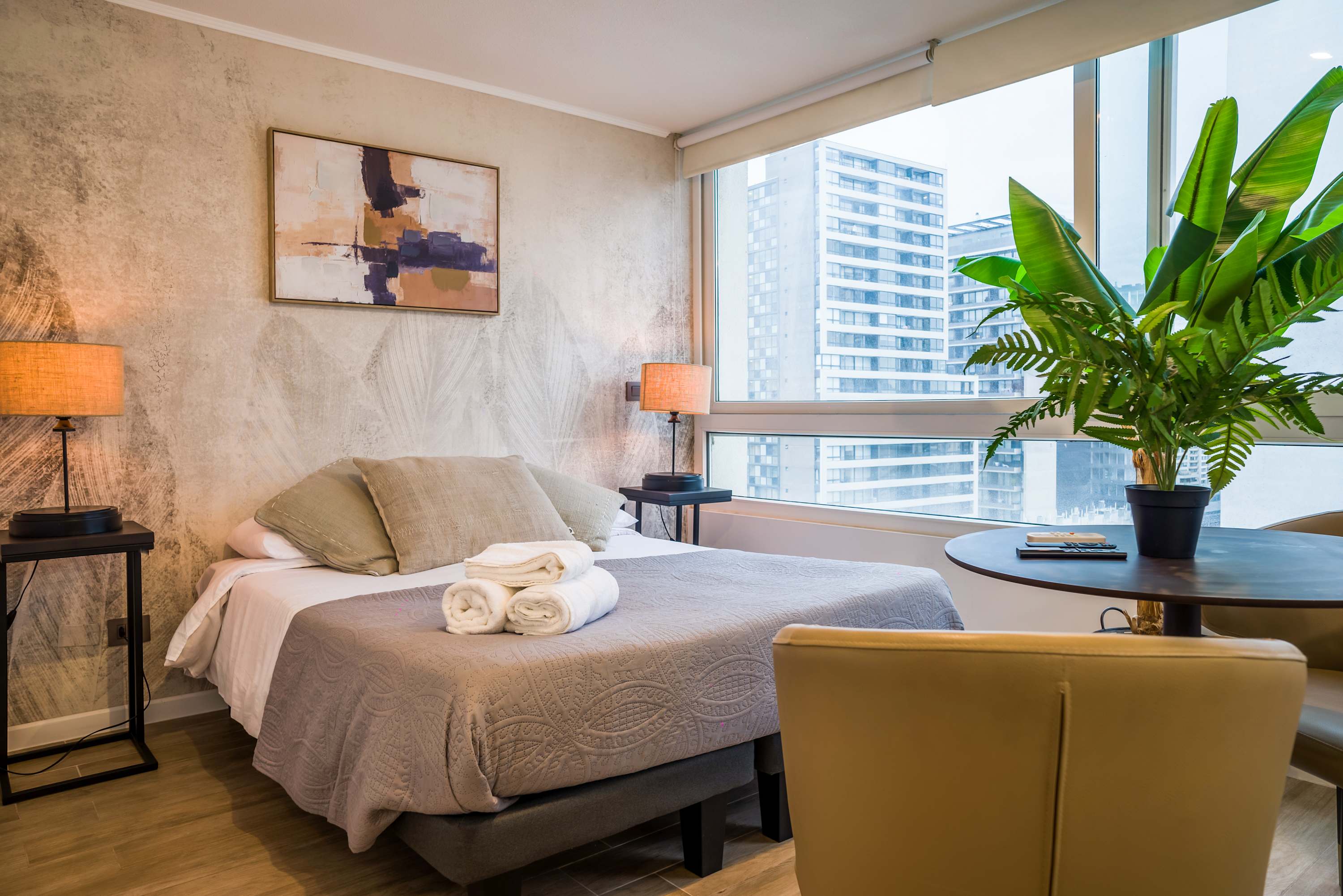 Studio bedroom with layered neutral bedding, folded towels, and expansive corner windows overlooking Santiago’s urban skyline.