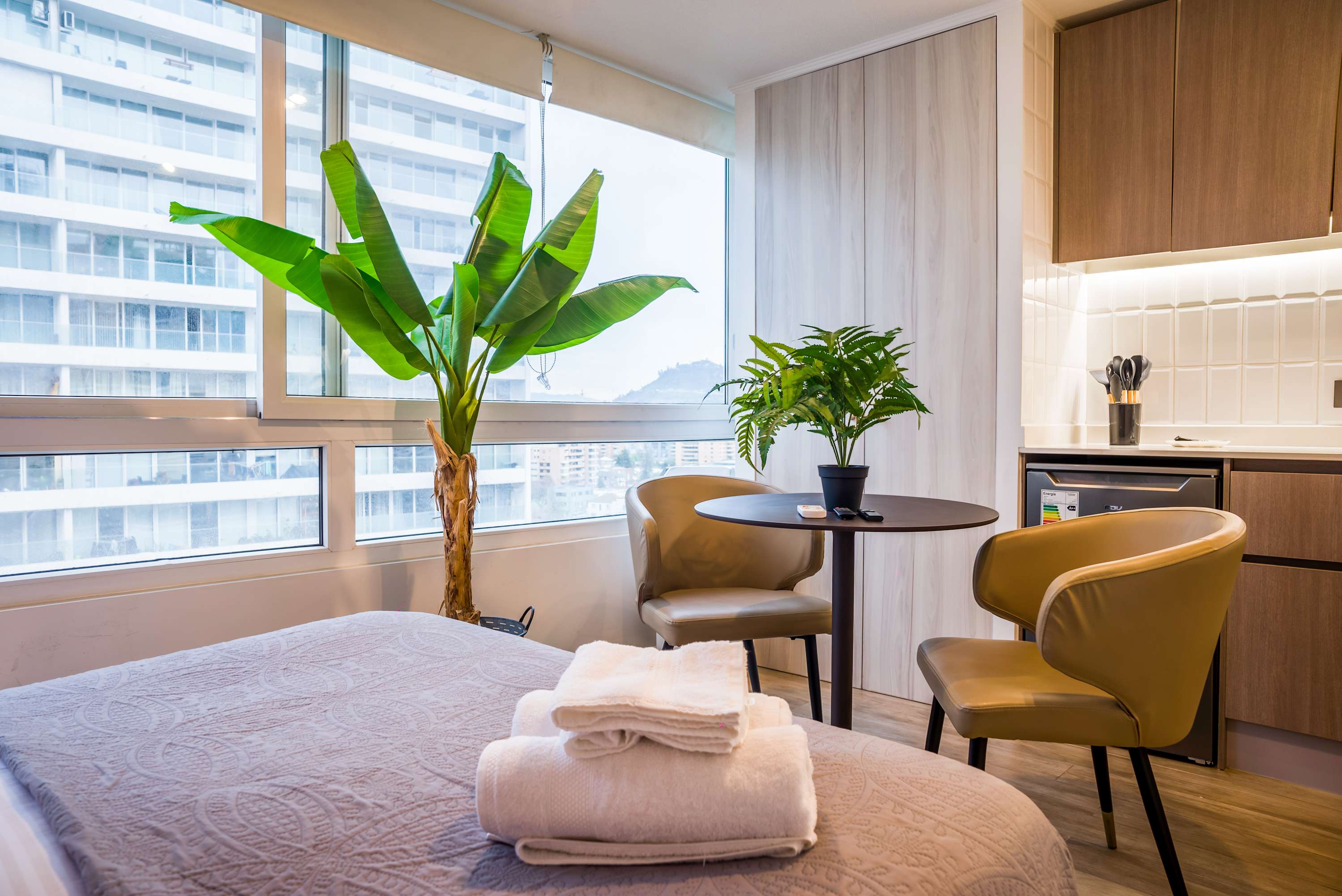 Bright studio kitchen and dining area with wood cabinetry, a round table for two, and expansive windows overlooking the city.
