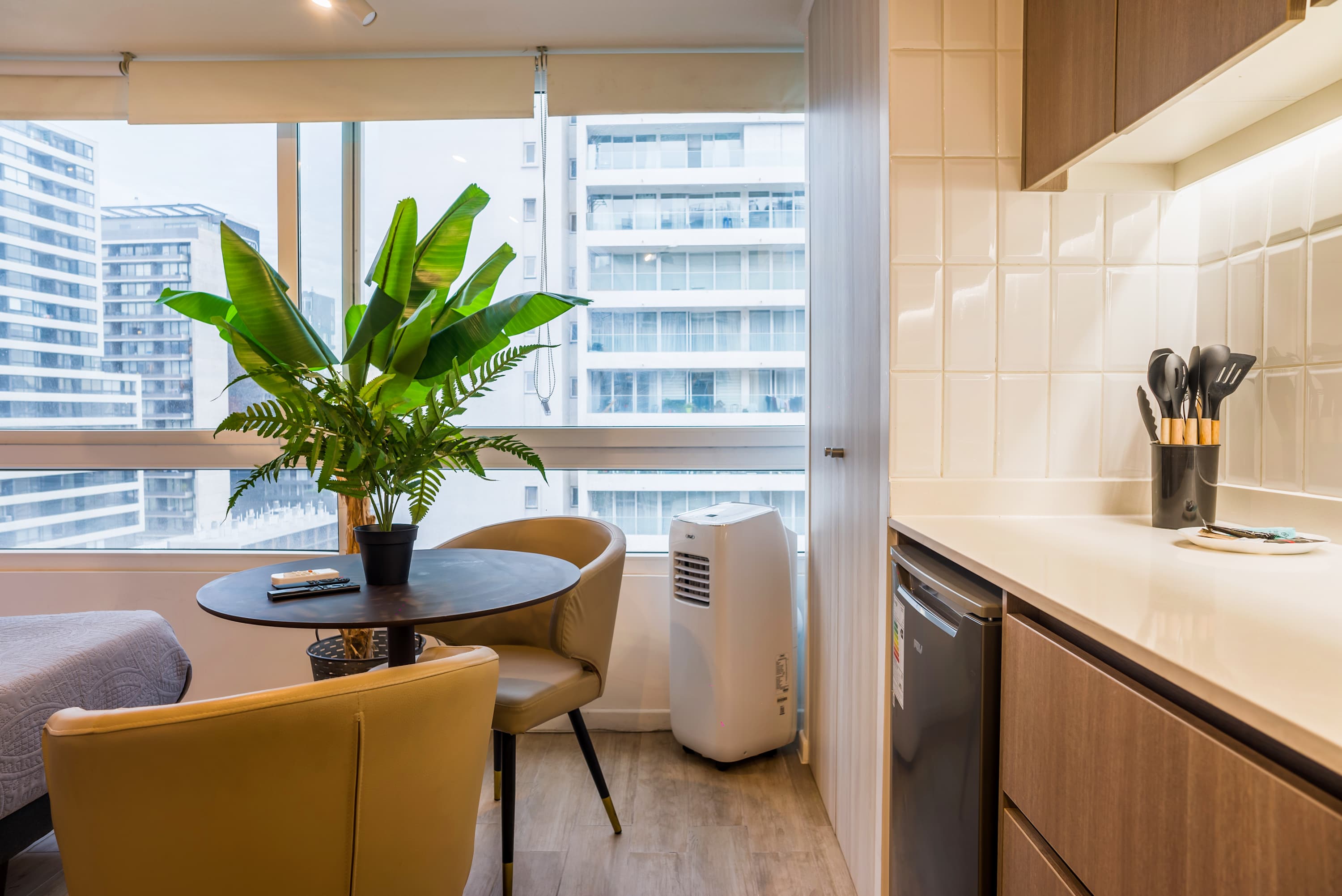 Bright studio living area with two armchairs, a round dining table, and leafy greenery beside expansive windows overlooking the city.
