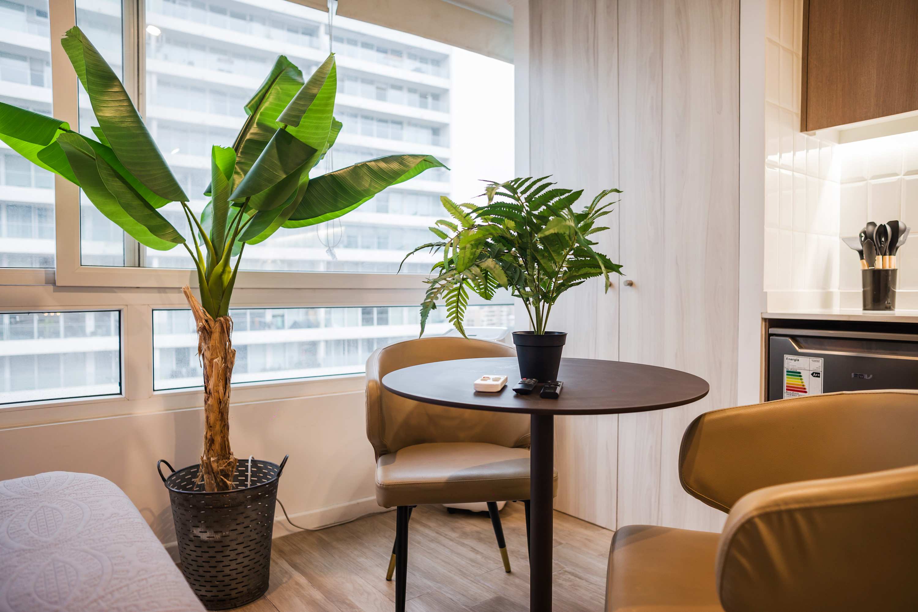 Studio dining nook with a round table, tan chairs, and bright floor-to-ceiling windows framed by lush green plants.