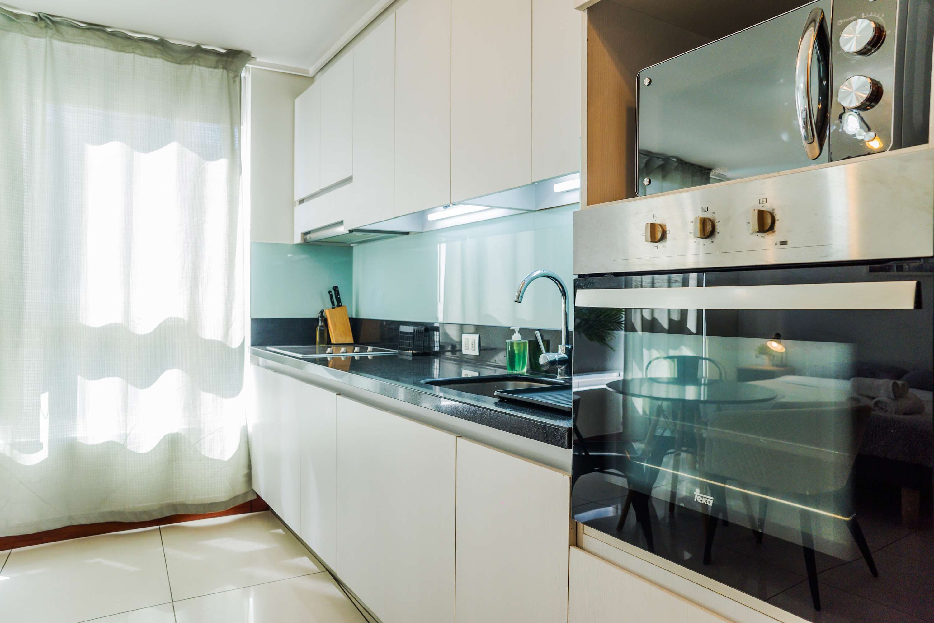 Private kitchen with sleek white cabinetry, induction cooktop, built-in appliances, and a bright window for natural daylight.