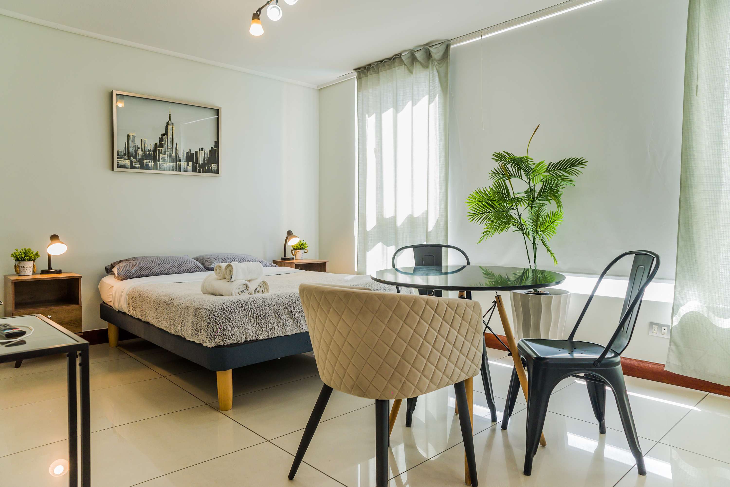 Bright studio living space with a neatly made bed, round glass dining table, sheer-curtained windows, and a tall green palm.