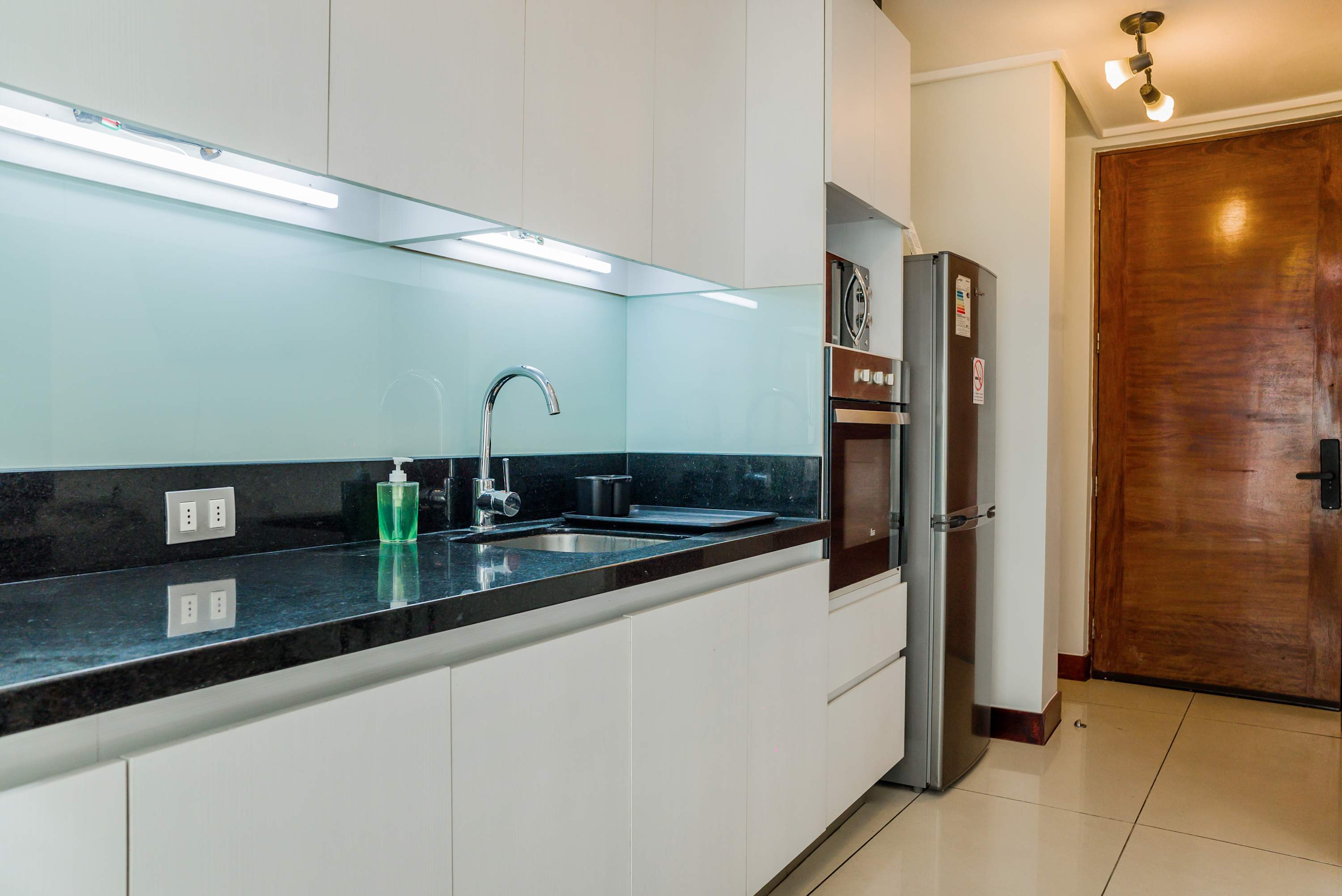 Private kitchen with sleek white cabinetry, polished black countertops, a chrome sink, built-in oven, and stainless-steel refrigerator.