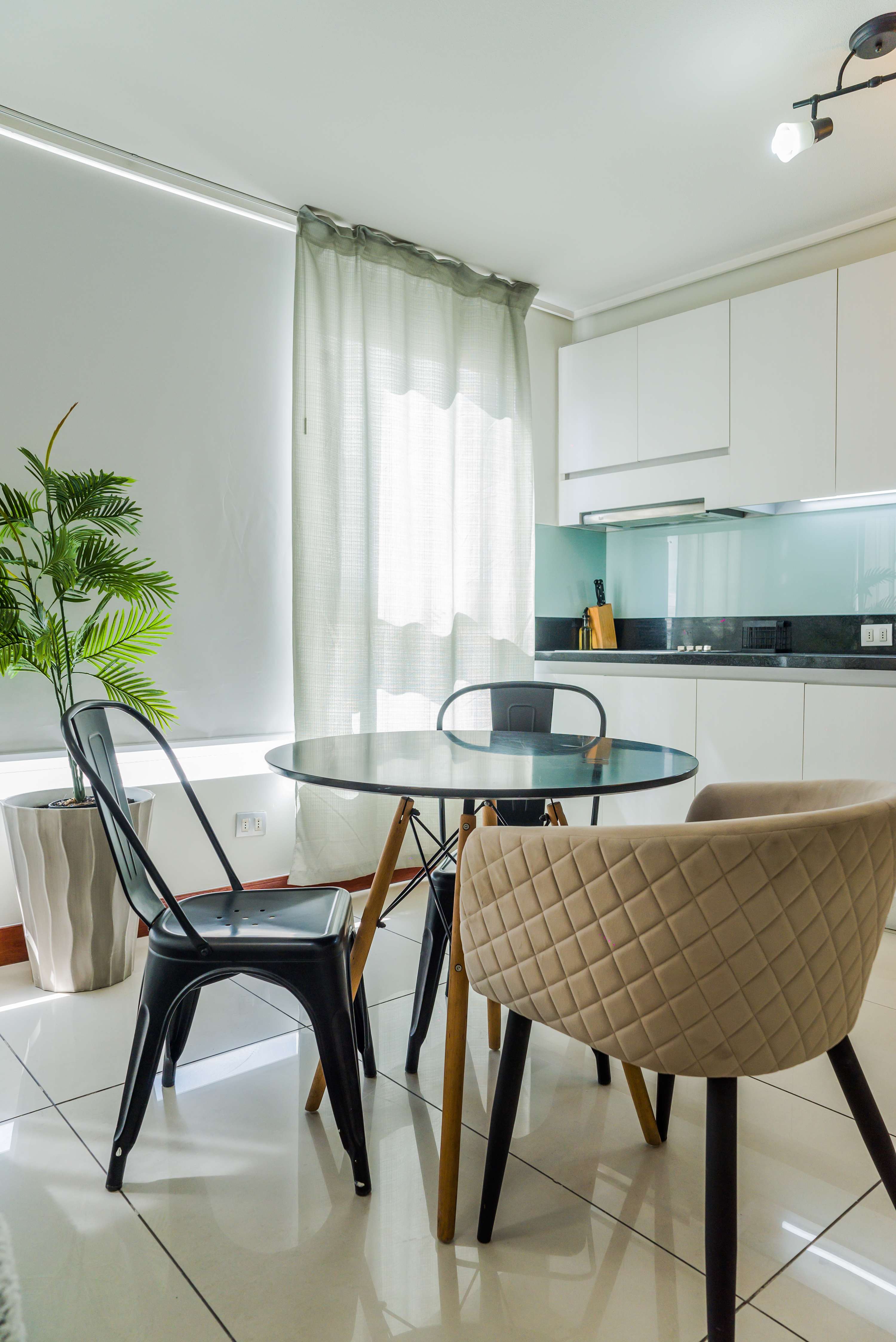 Private dining area with a round glass table, modern white kitchen, and soft daylight filtered through sheer curtains.
