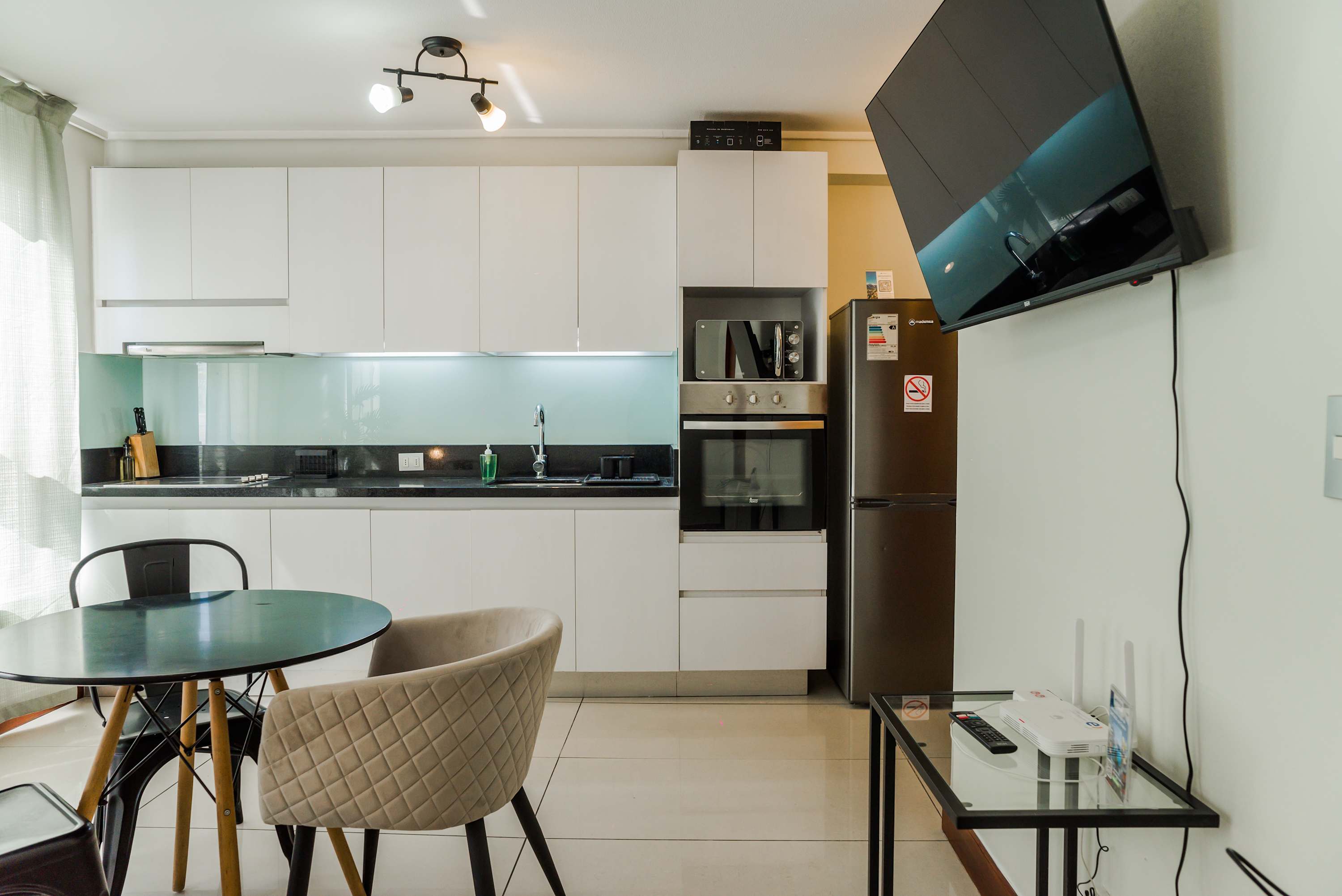 Private studio kitchen with sleek white cabinetry, a turquoise glass backsplash, full-size refrigerator, built-in oven, microwave, and a dining table for two.