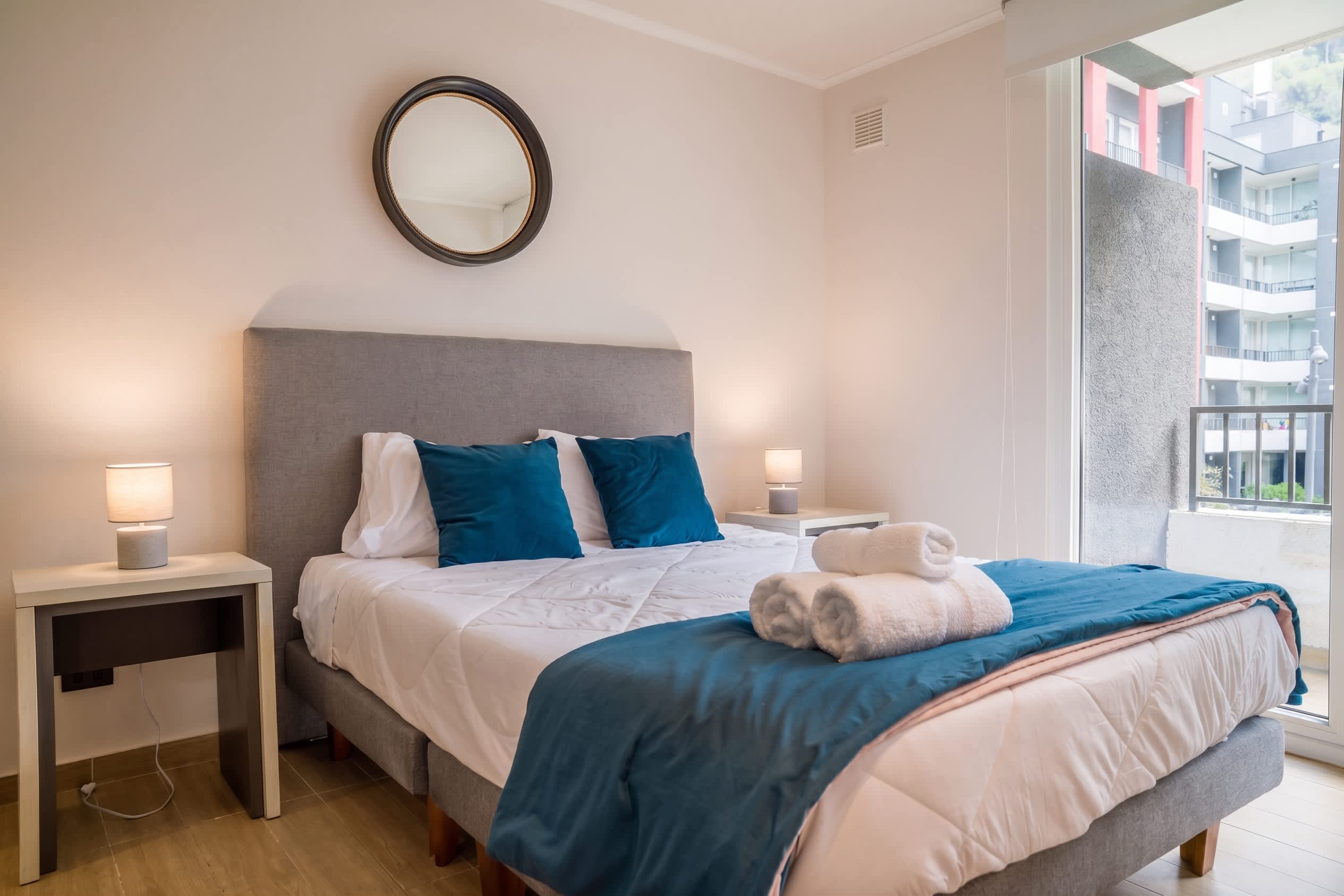 Comfortable bedroom with a gray upholstered bed, teal accents, bedside lamps, and bright balcony access overlooking the surrounding apartments.