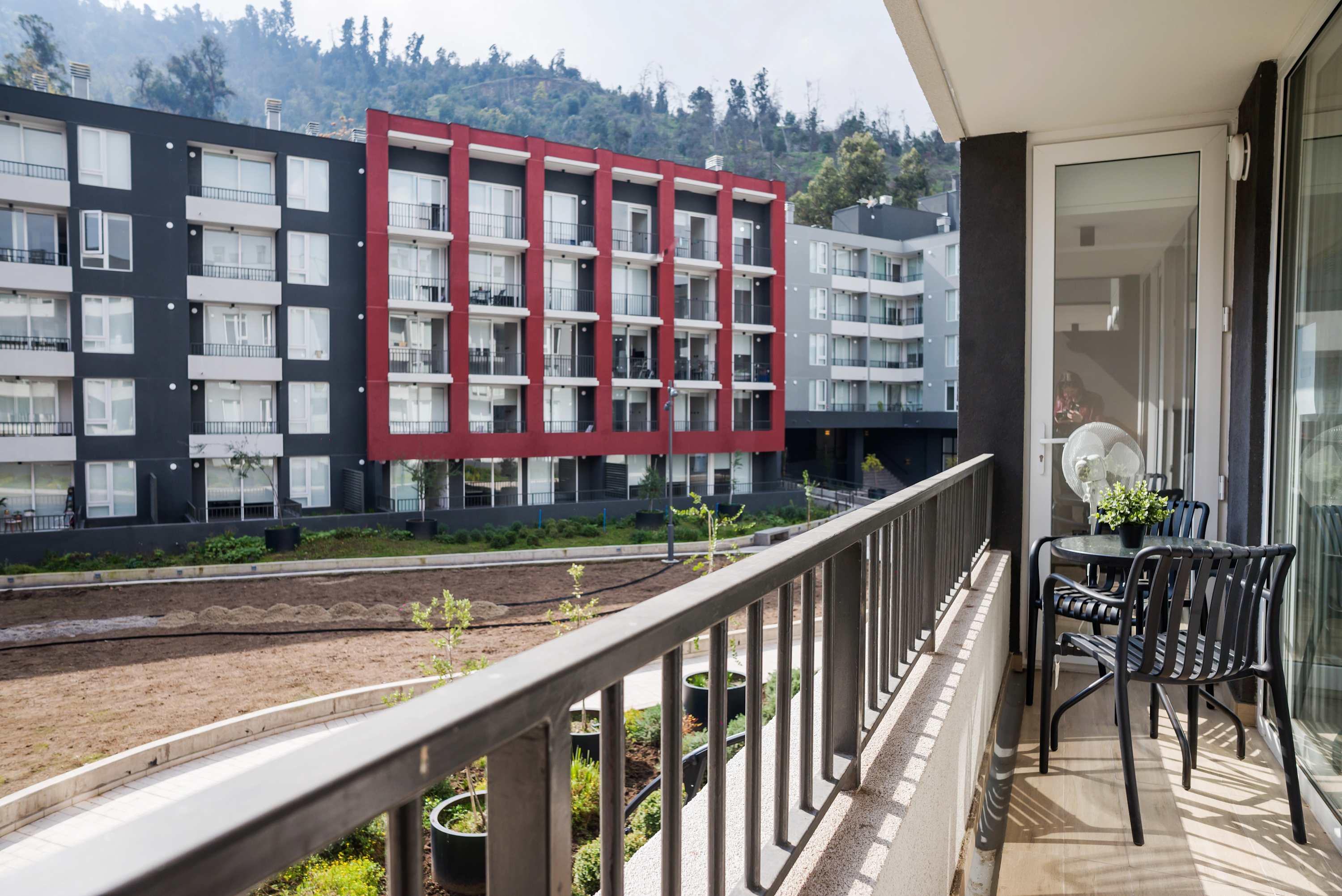 Private balcony with a four-seat café table, potted greenery, and a wooded hillside view beyond the modern apartment complex.